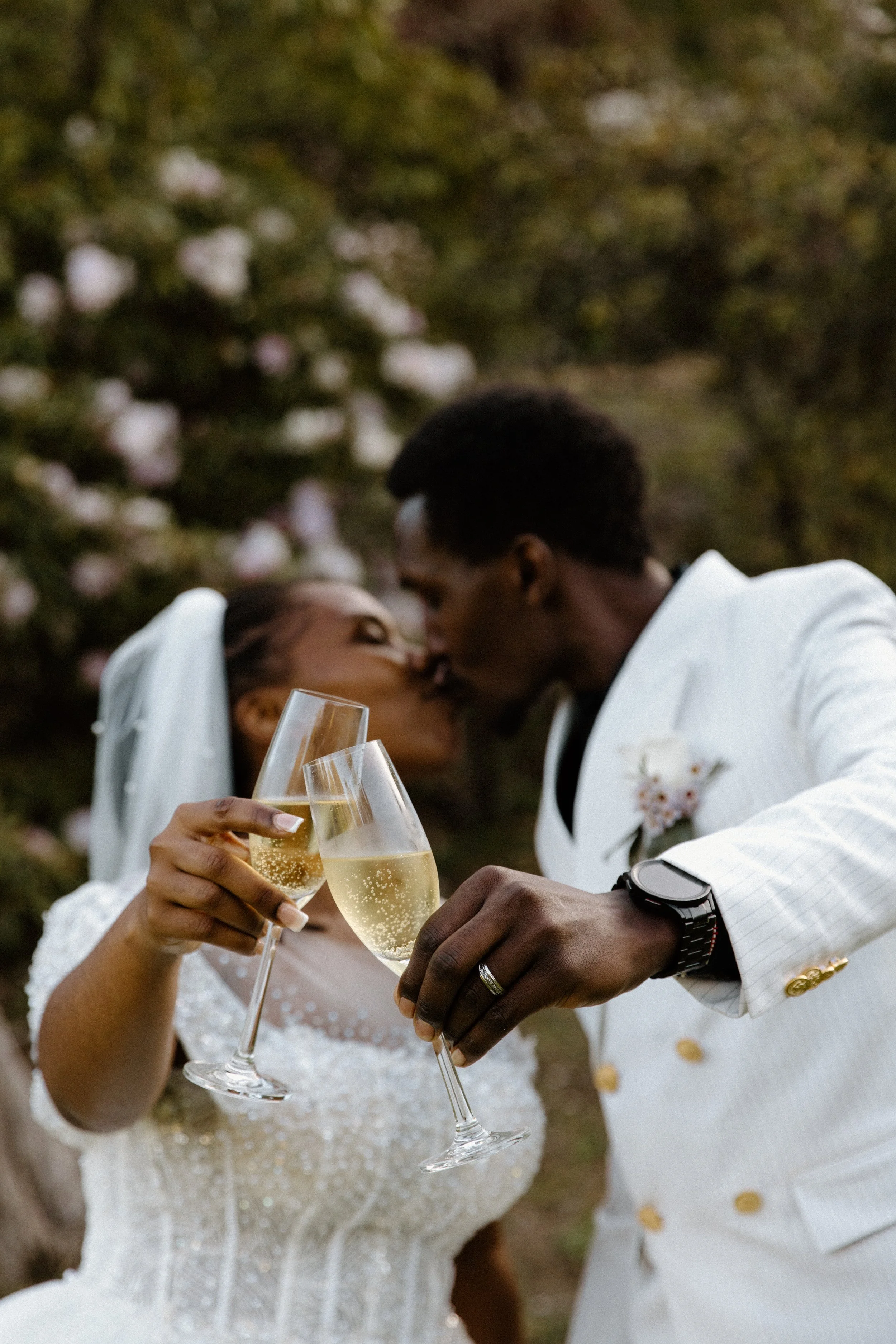 A newlywed couple in wedding attire kissing while holding champagne glasses outdoors.