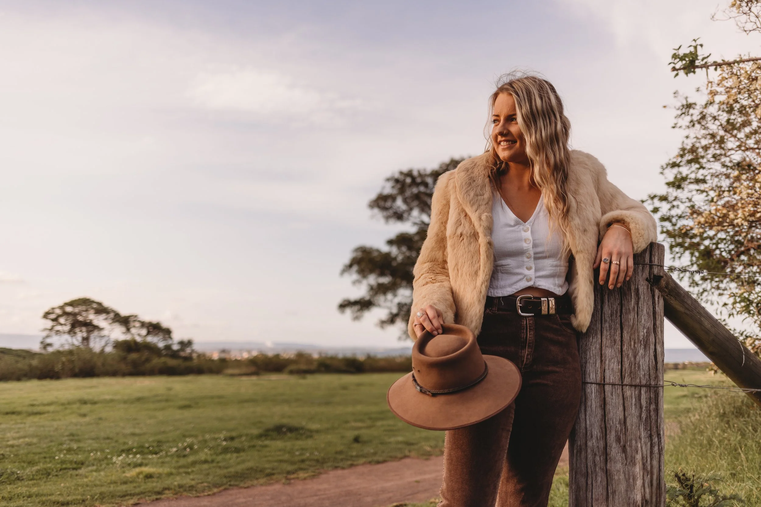 A woman standing outdoors on a grassy field, leaning against a wooden post, holding a hat in her right hand, wearing a beige fur coat, white shirt, and brown pants, smiling and looking away from the camera