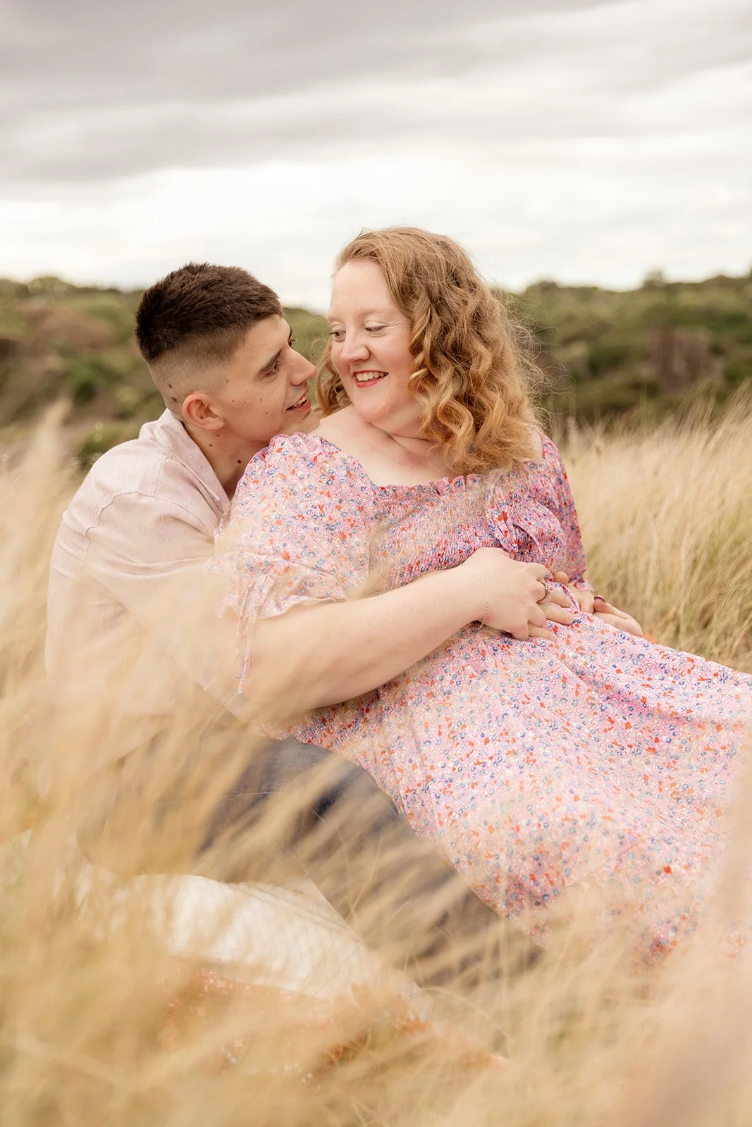 A young man and a pregnant woman sitting together in a field of tall grass, with the man looking affectionately at the woman who is smiling and holding her belly, under an overcast sky.
