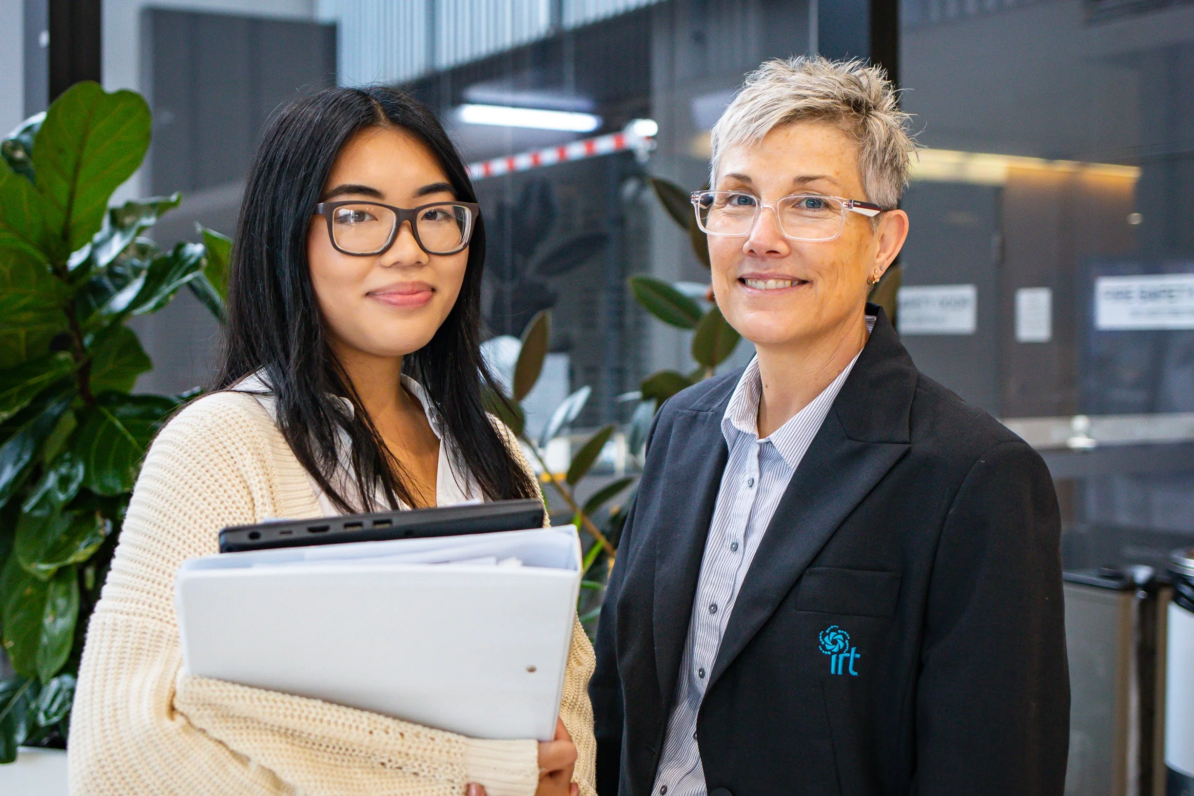 Two women standing indoors, one holding files and the other smiling at the camera. Behind them are green plants and dark-colored walls.