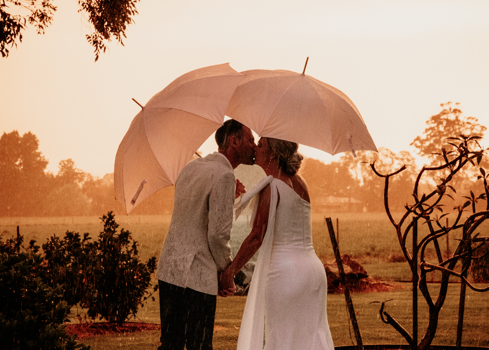 A couple in wedding attire shares a kiss under pink umbrellas in a garden at sunset.