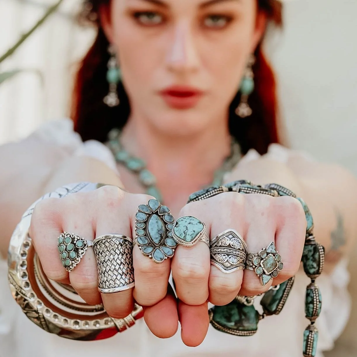 A woman with red hair and blue eyes displays her hand adorned with multiple silver rings with turquoise stones, wearing jewelry and a bohemian style outfit.