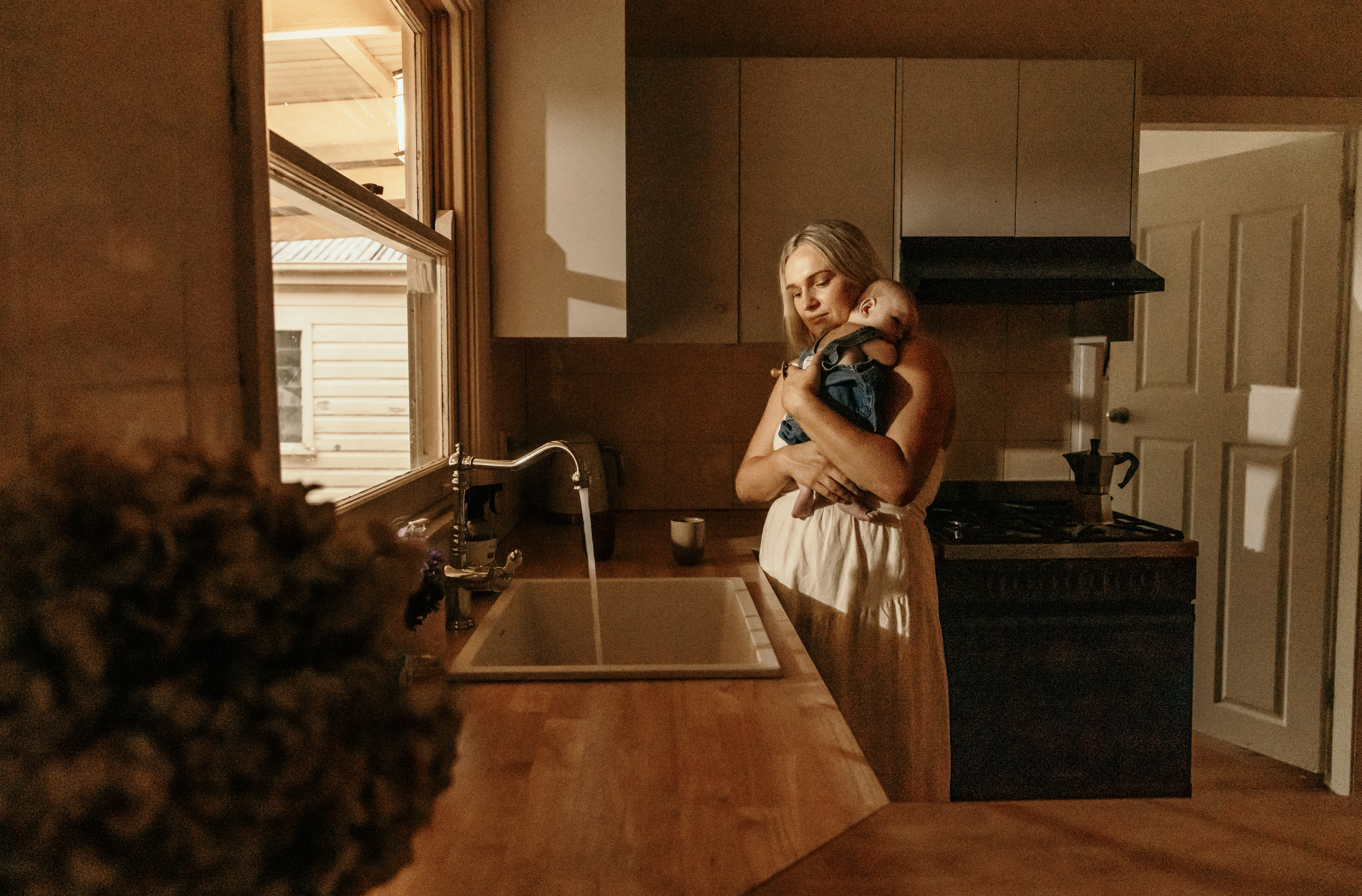 A woman holding a baby in a kitchen with warm lighting and wooden cabinets.