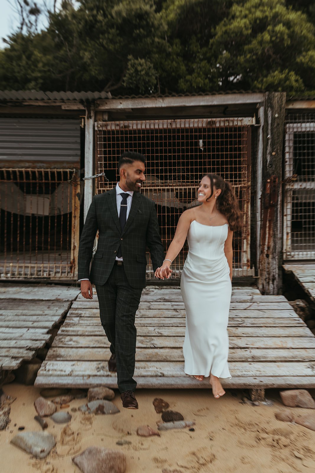 A newlywed couple holding hands and walking on a wooden dock in Sandon Point, smiling and looking at each other. The man is dressed in a black suit, and the woman is wearing a white wedding dress, with an outdoor background.