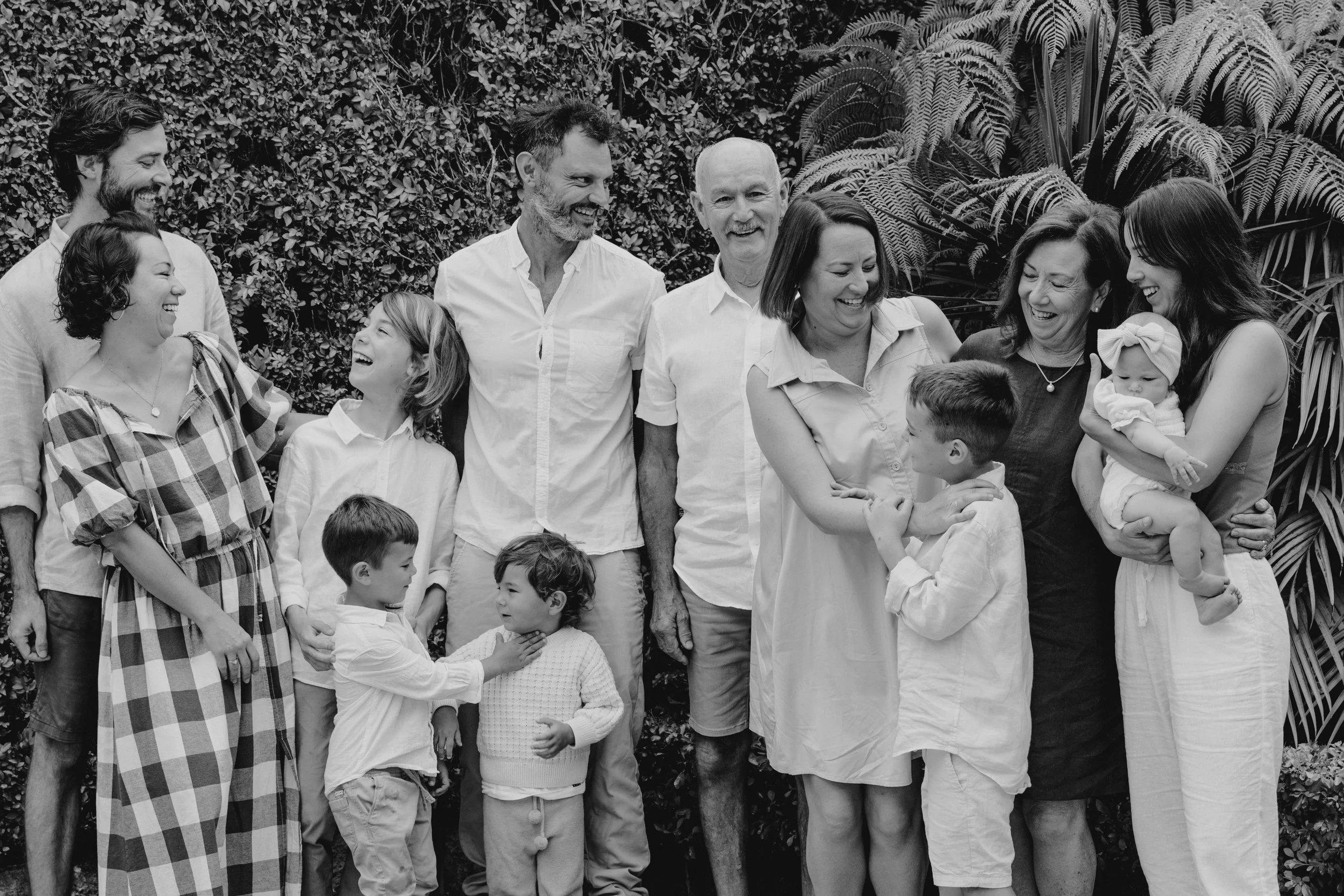 A black and white photo of a joyful, multi-generational family gathering outdoors, with people laughing and interacting against a leafy background.