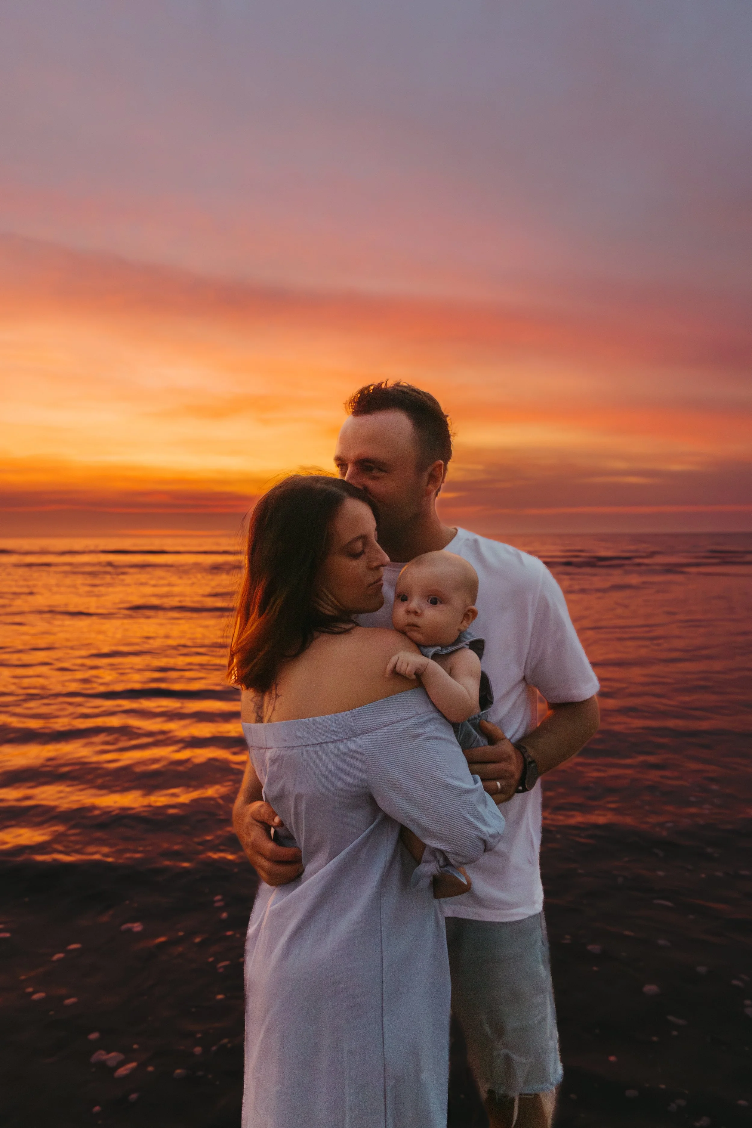 A family of three, consisting of a woman, a man, and a baby, standing on the beach during a colorful sunset. The woman is holding the baby, and the man is gently embracing both, with the ocean in the background.