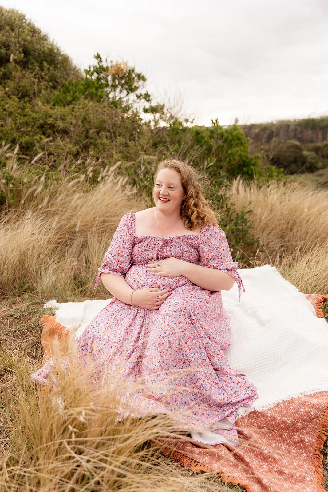 A woman with curly hair wearing a floral dress, sitting on blankets in a grassy field, smiling and holding her stomach.