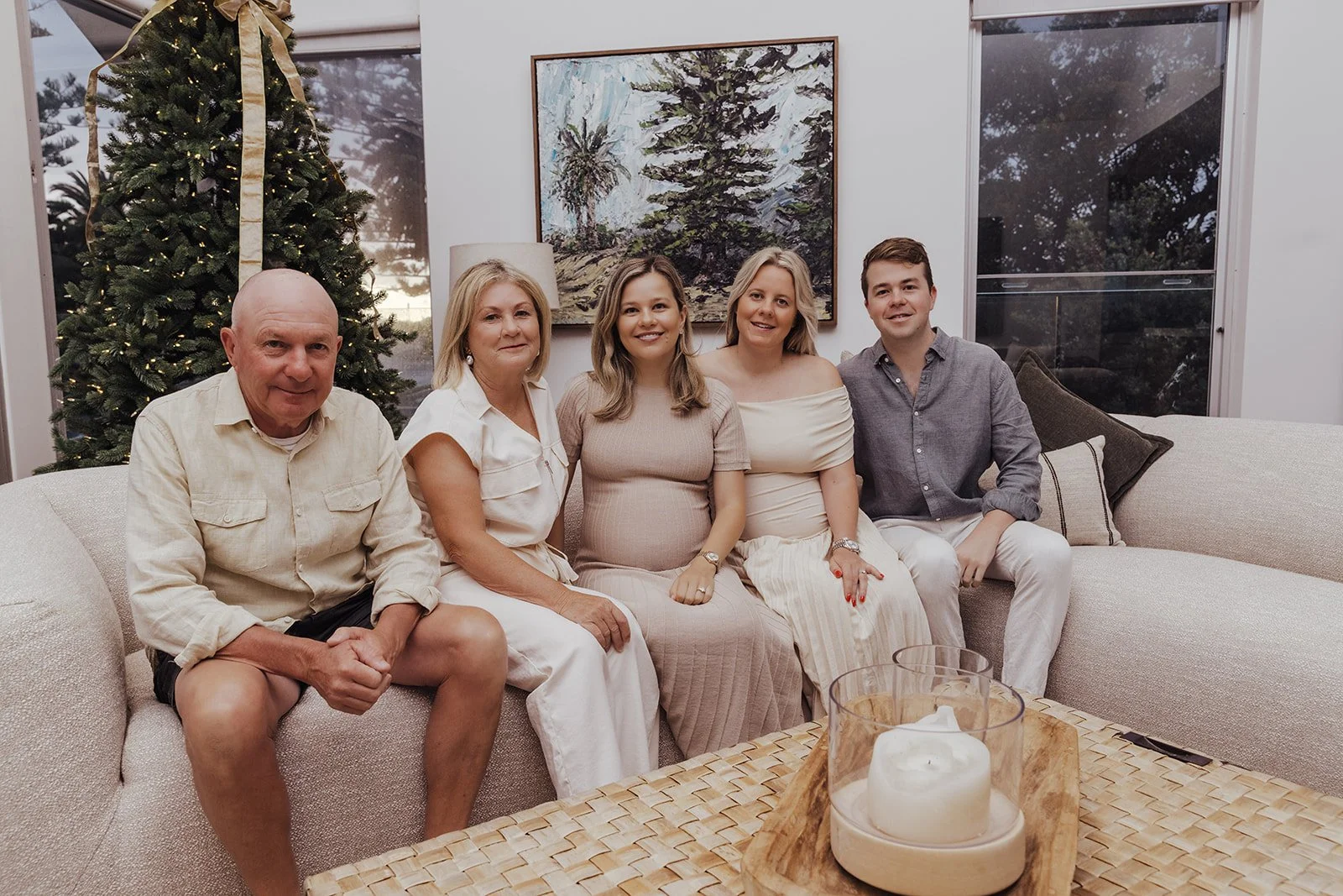 A family of five sitting on a beige sofa in a living room, decorated for Christmas with a Christmas tree and framed artwork, during the daytime.