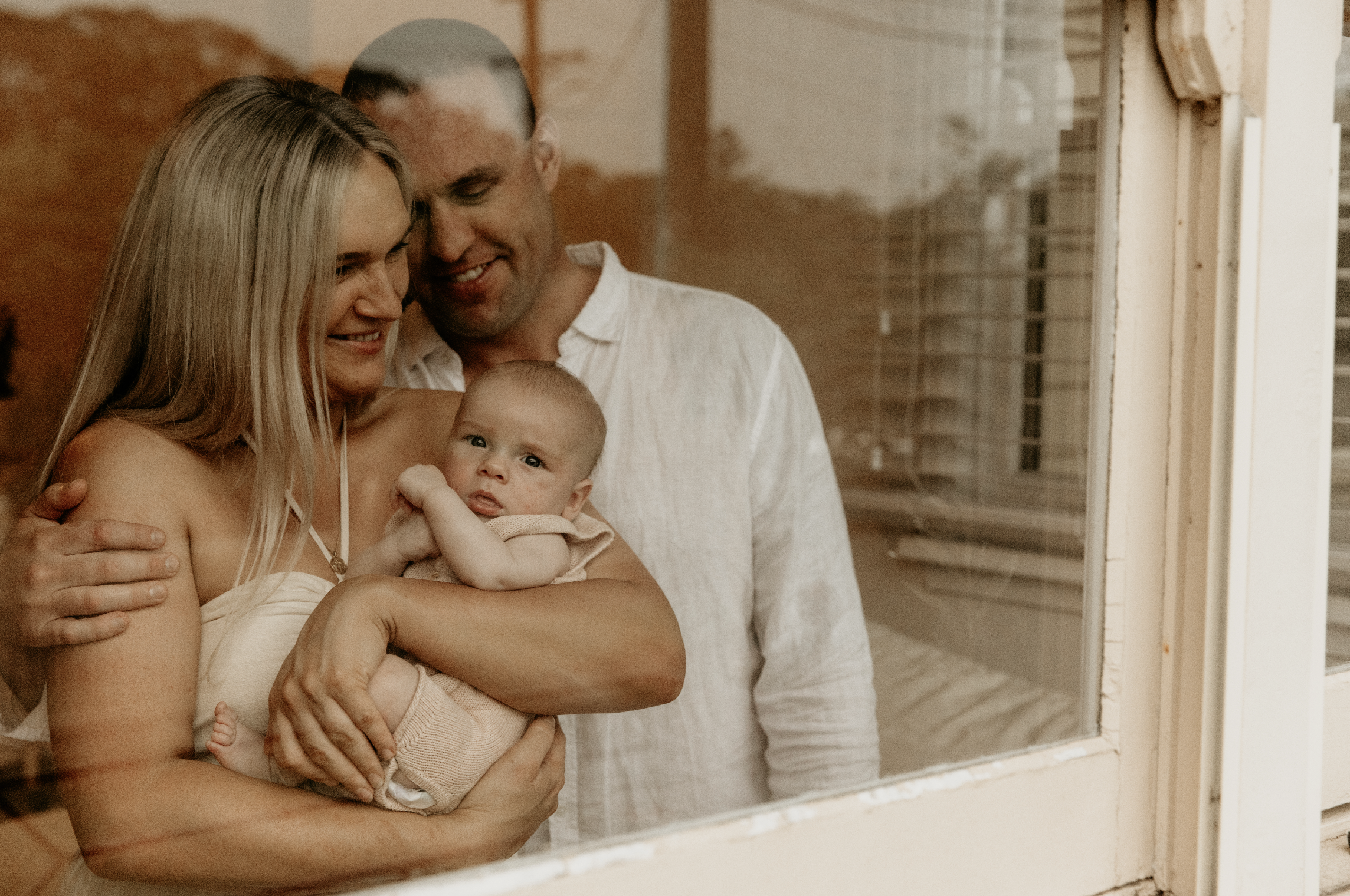 A family of three through a window: a woman holding a baby, and a man standing behind them, all smiling.