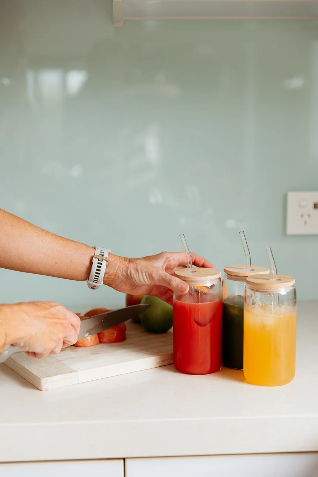 Person pouring colorful juice from jars with straws on a kitchen countertop, with a cutting board and fruit in the background.