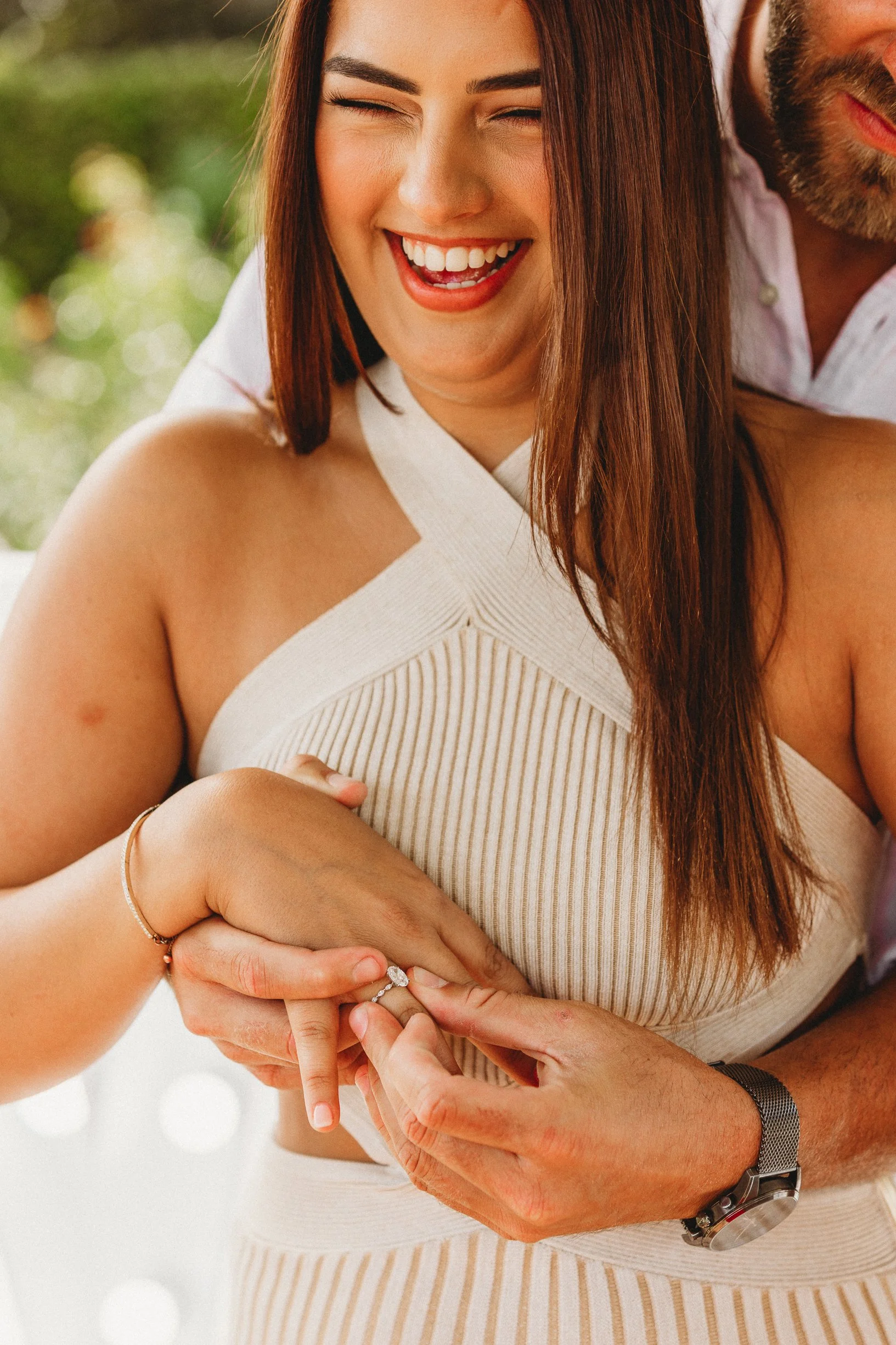 A woman with long brown hair and a white sleeveless dress is smiling, showing her teeth, while a man behind her is placing a ring on her finger.