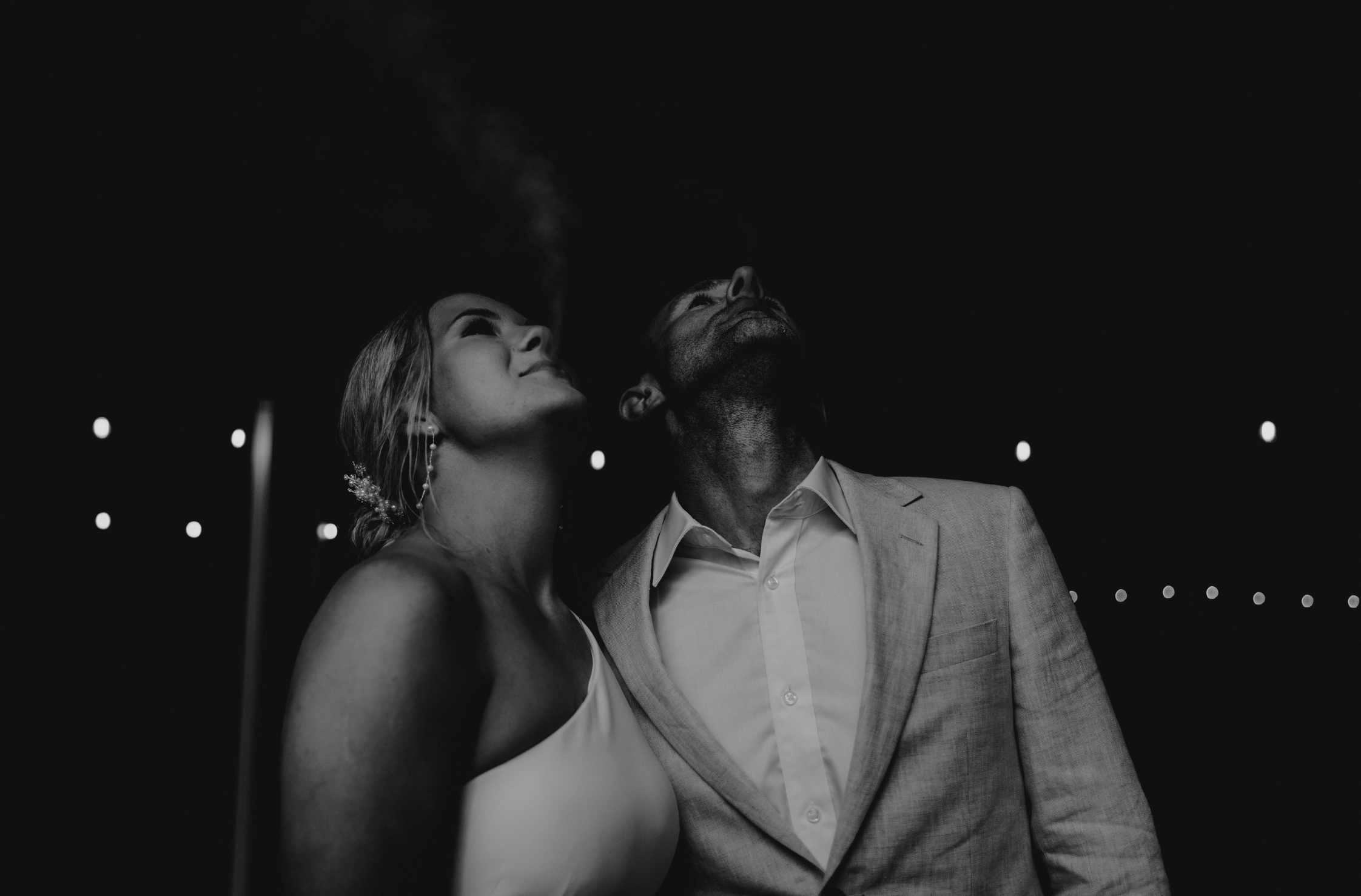 Black and white photograph of a man and woman looking upward with smoke rising from the woman’s mouth, dressed in formal attire, under string lights at night.