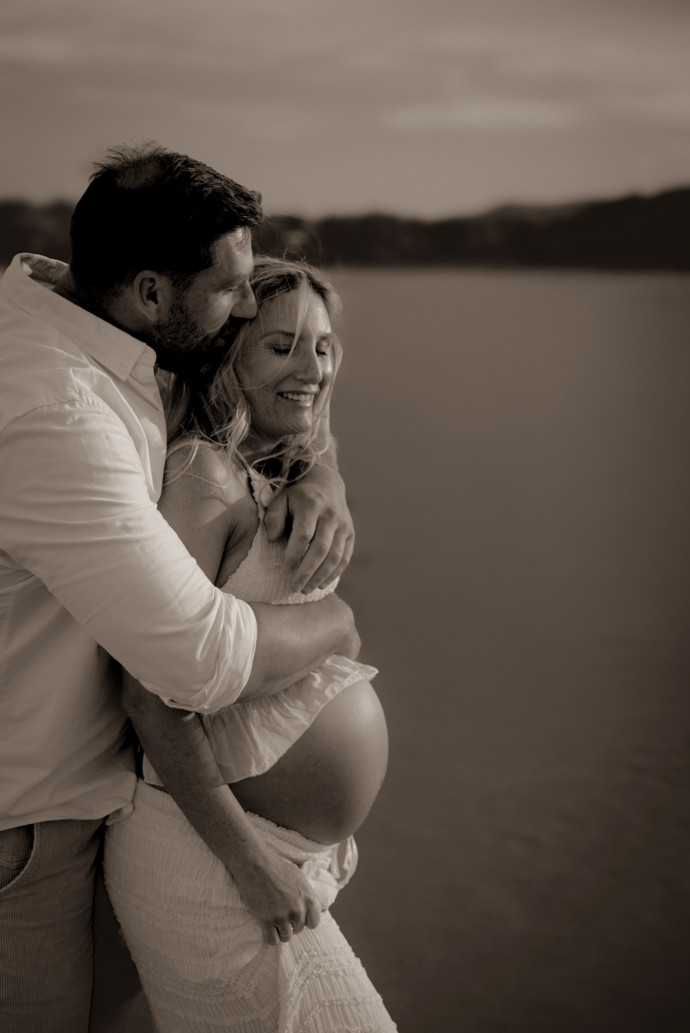A close-up black-and-white photo of a pregnant woman in a dress smiling as a man embraces her from behind near a body of water at sunset.
