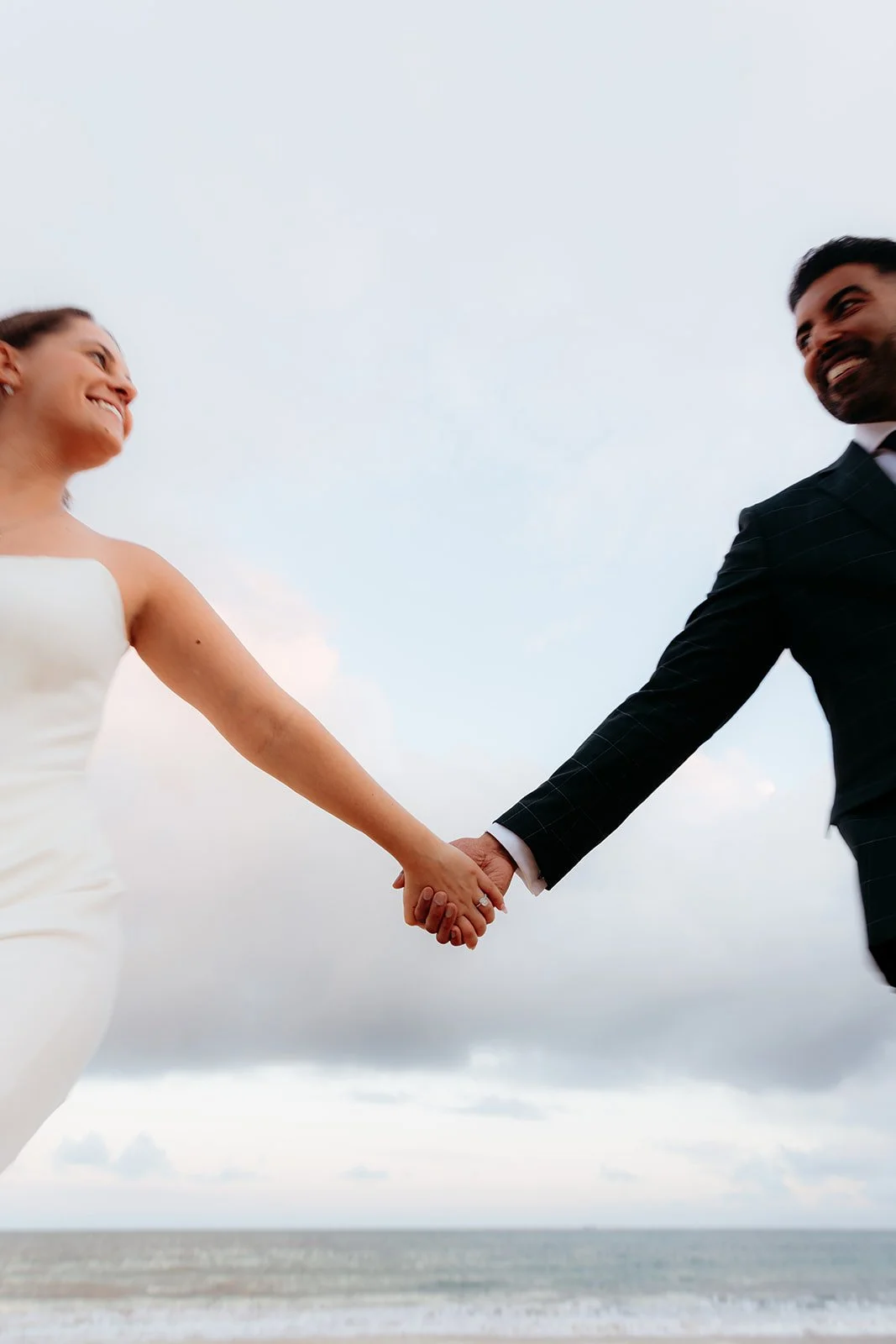 A bride and groom holding hands and smiling at each other on the beach during their wedding. Vintage film