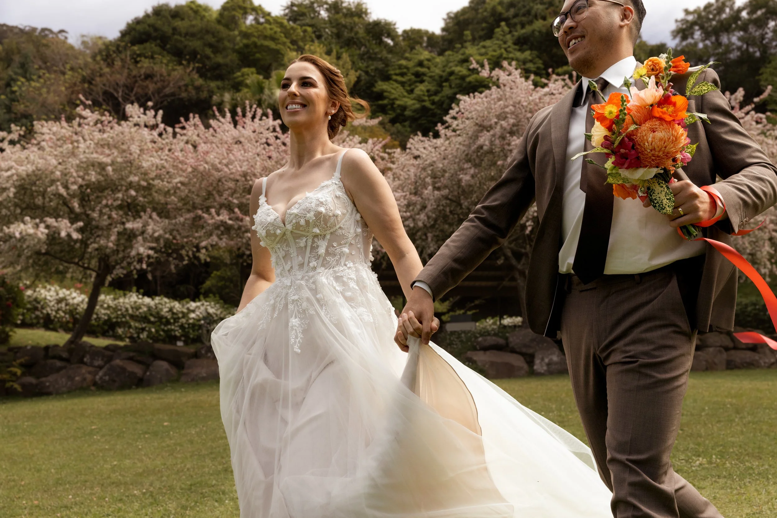 A bride and groom holding hands and walking outdoors in a park with blooming pink cherry blossom trees in the background.