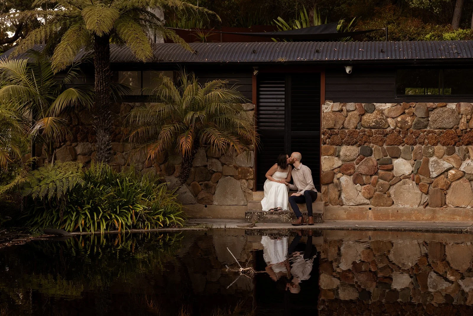 A couple sitting on a porch by a pond, sharing a kiss. The woman is wearing a white dress, and the man a light shirt and dark pants. The scene is surrounded by lush greenery and a stone building.