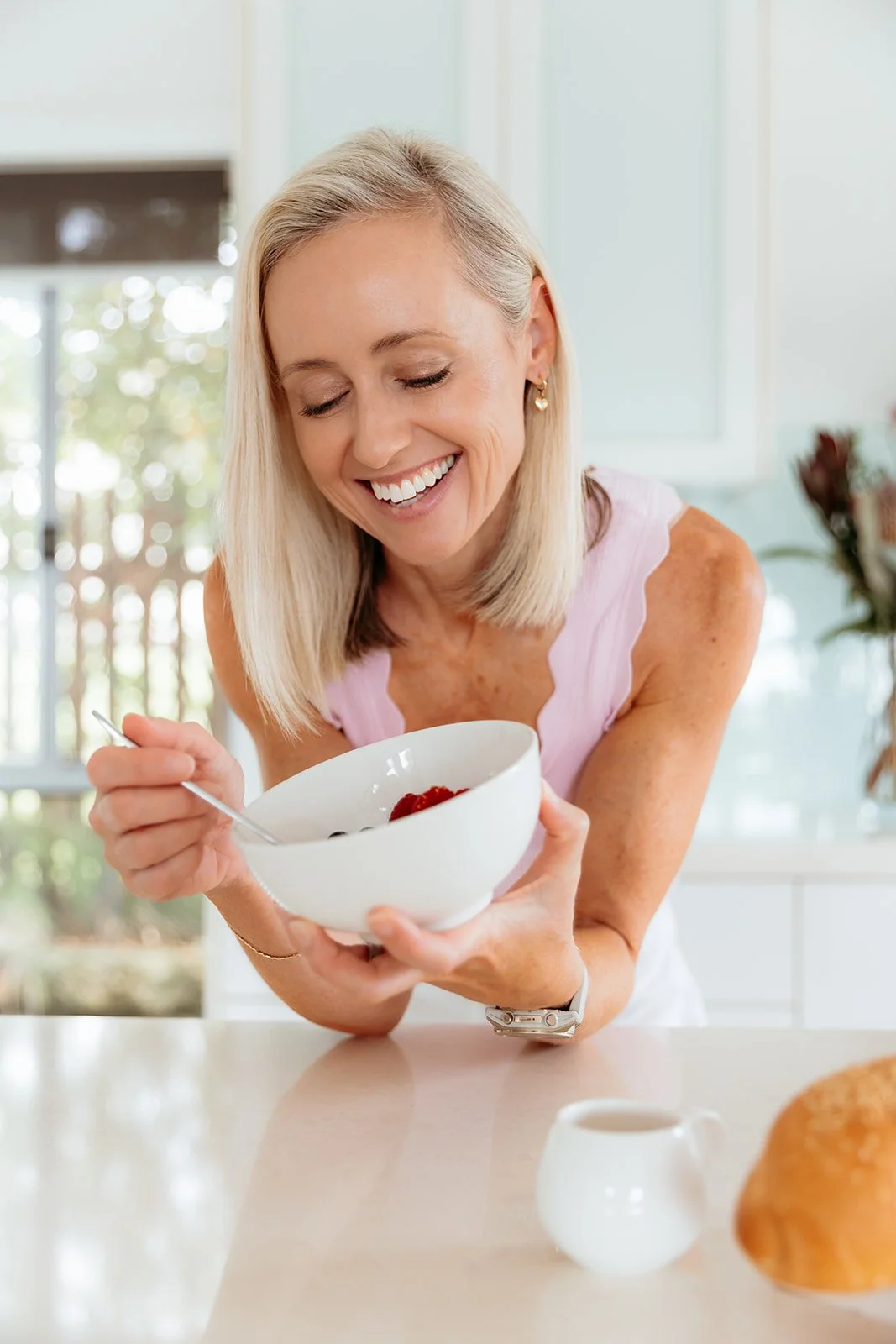 A smiling woman in a pink sleeveless top holding a white bowl filled with berries in a sunny kitchen.