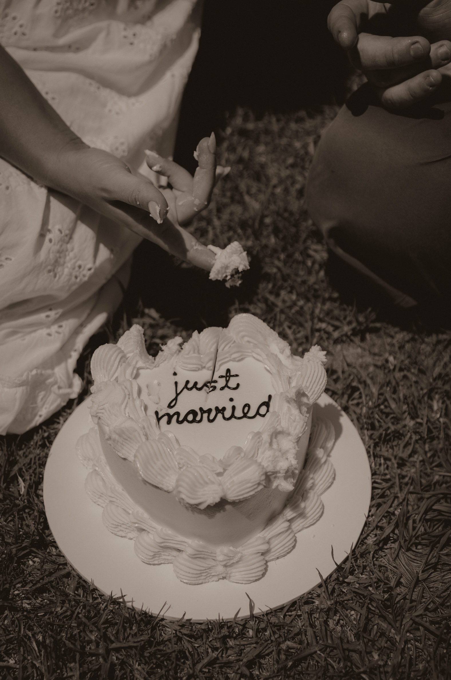 A wedding cake with the words 'just married' written on it, placed on the grass, with a person reaching towards it, possibly to cut or serve the cake.