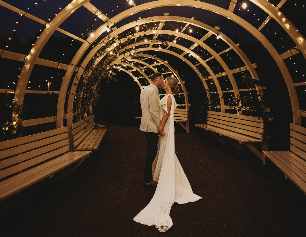 A newlywed couple stands close together under a decorative arch with string lights at night, holding hands and about to kiss.