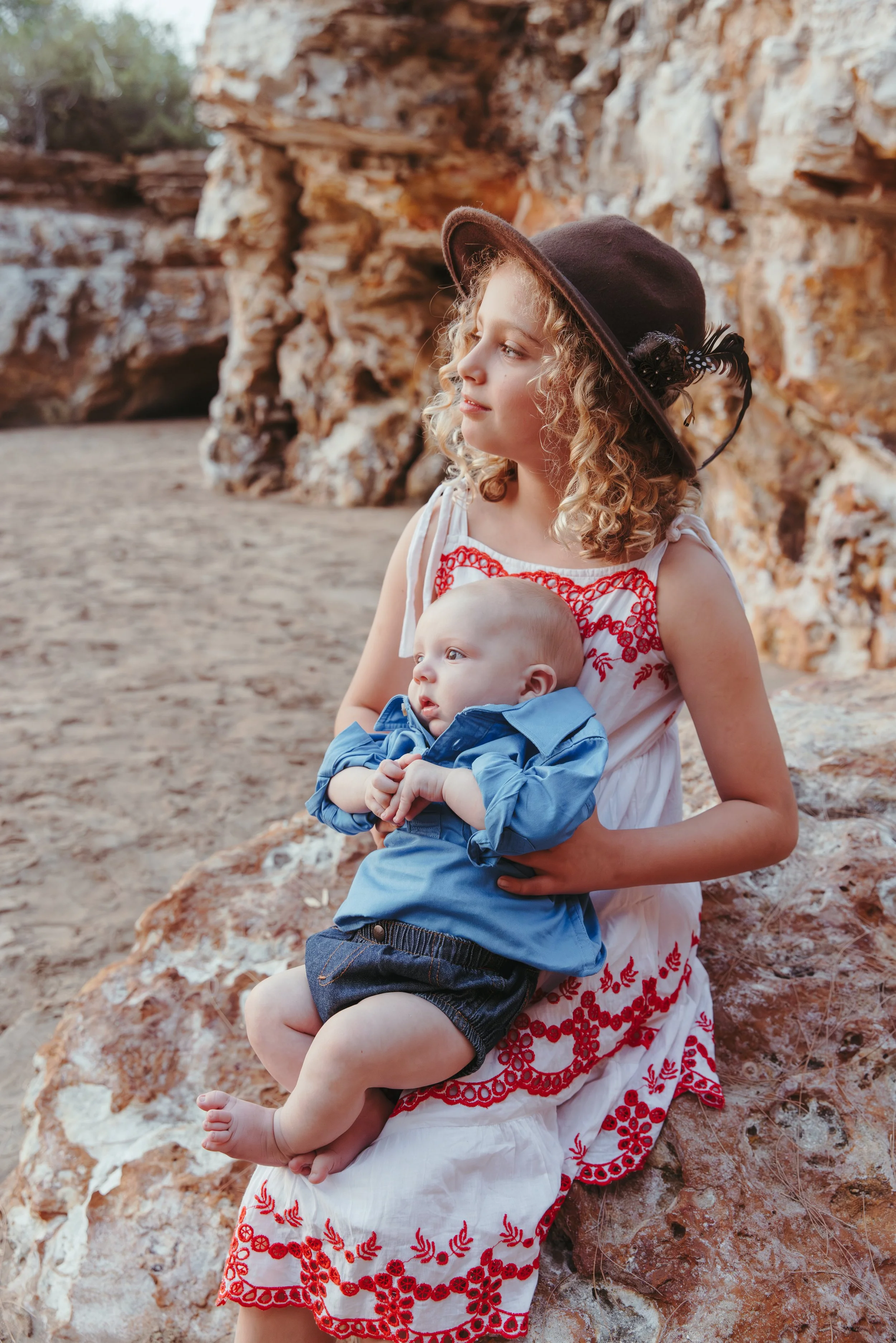 A young girl with curly blonde hair, wearing a large brown hat with a feather, sits outdoors on a rock holding a baby boy in her lap. The girl is dressed in a white dress with red embroidery, and the baby is wearing a blue shirt and shorts. Rocks and