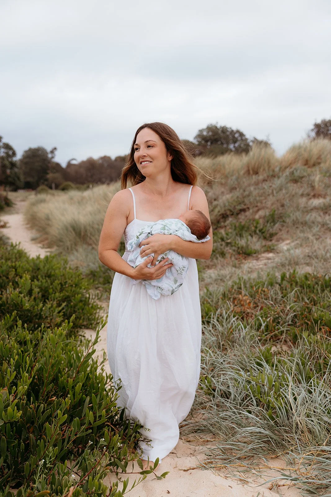 A woman in a white dress walking along a sandy path holding a baby wrapped in a blanket.