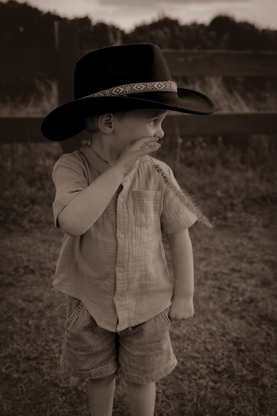 A young boy wearing a large black hat and a short-sleeved button-up shirt, standing outdoors at dusk or dawn, with a rural landscape and a wooden fence in the background.