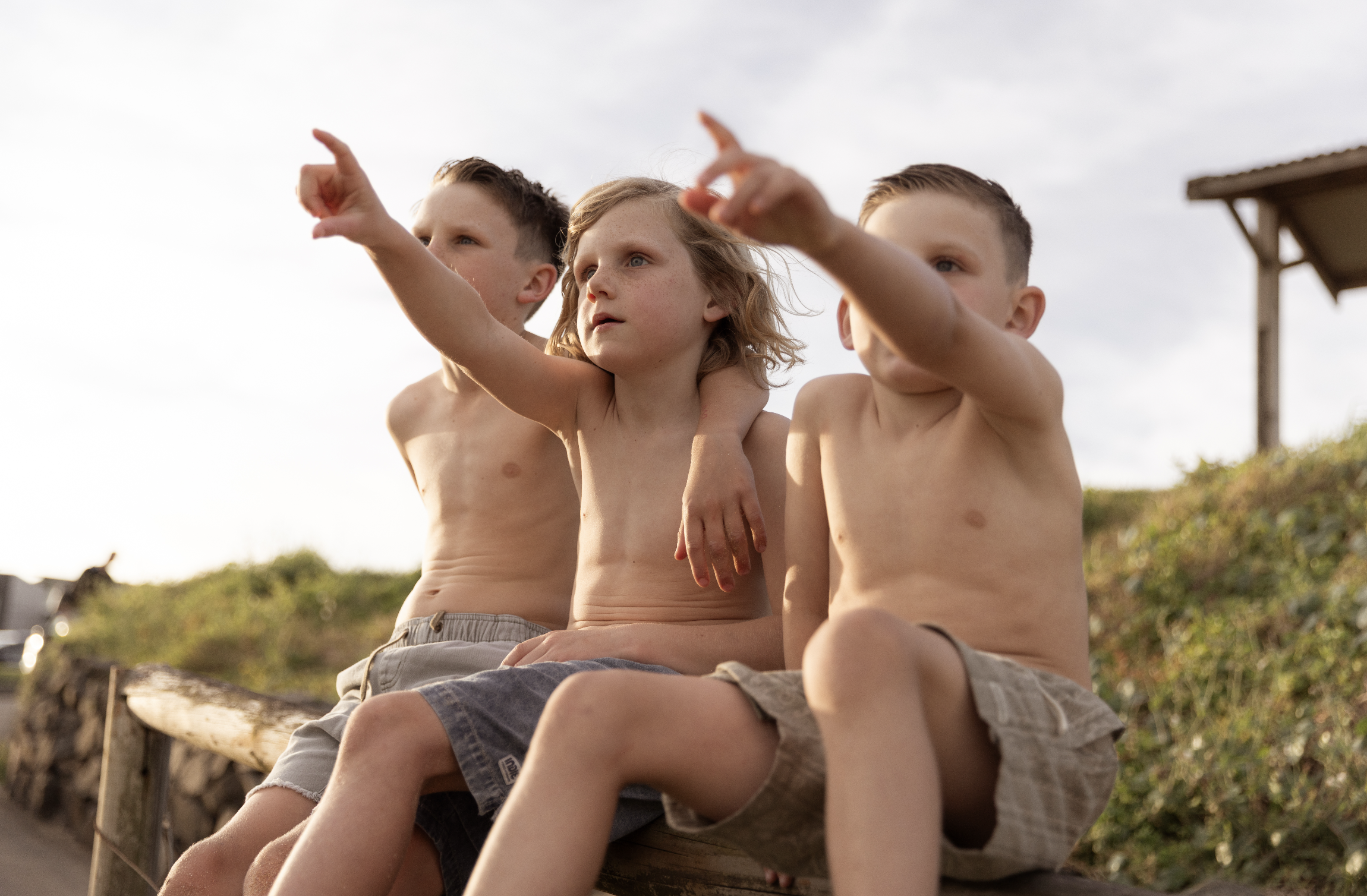 Three shirtless children with shorts sitting on a wooden bench outdoors, pointing at something in the distance during daytime.