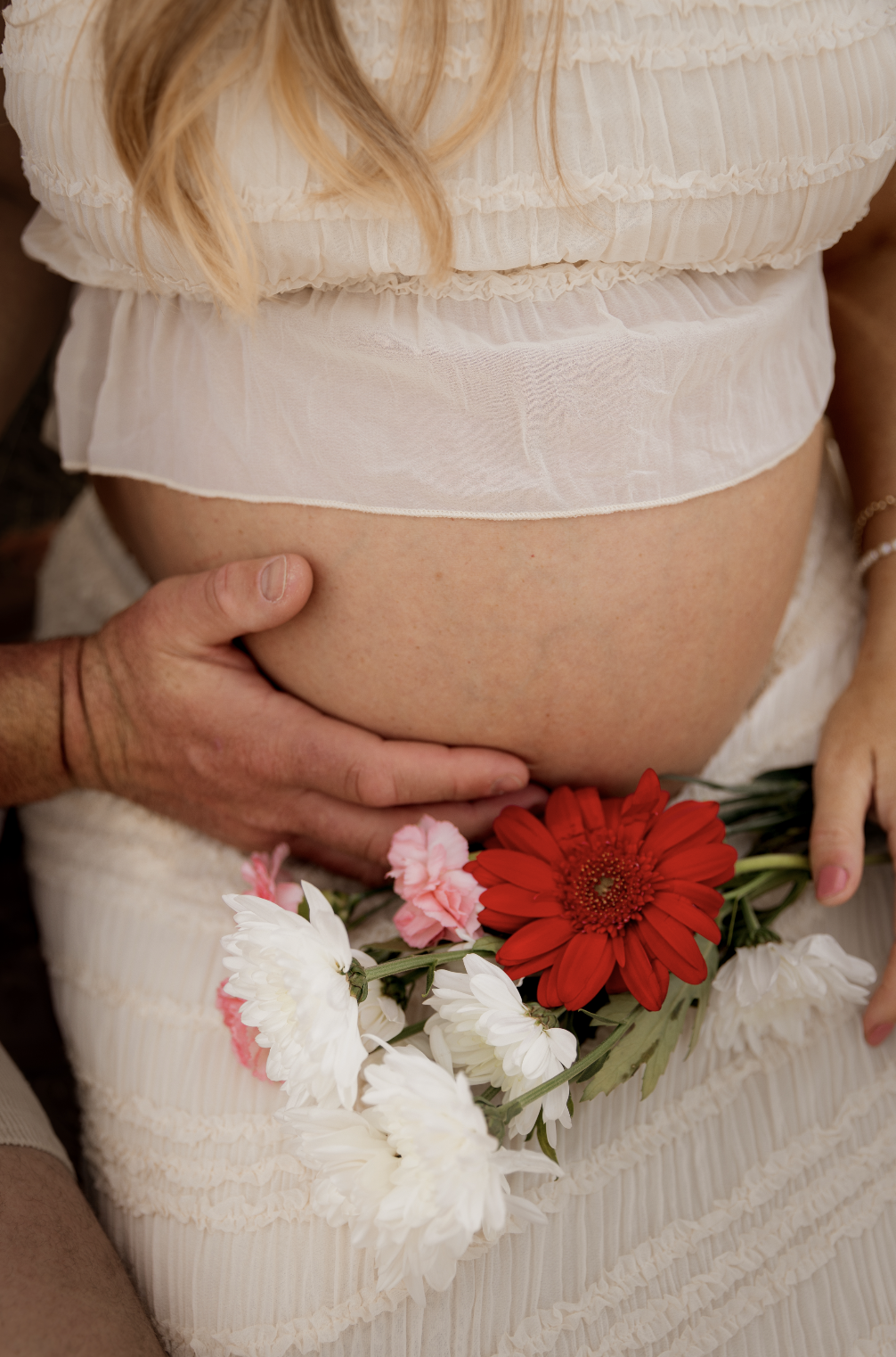 A pregnant woman wearing a light, flowy, ruffled dress holding a bouquet of red, white, and pink flowers with a hand gently resting on her belly