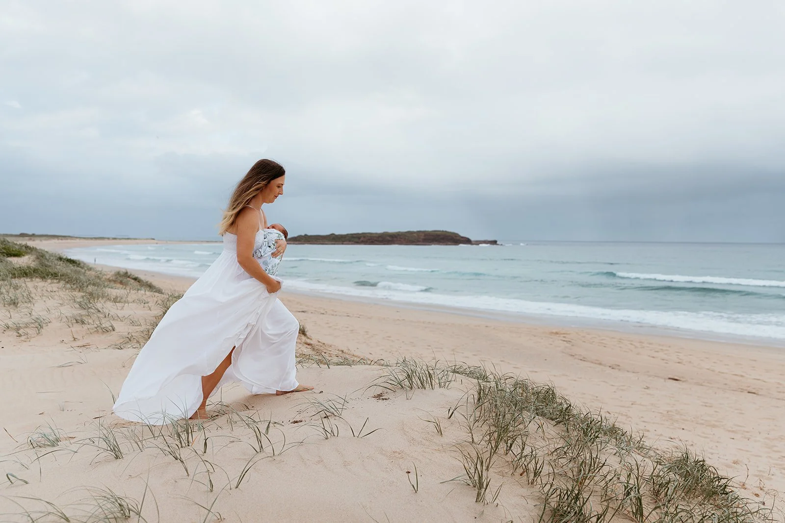 A woman in a white dress on the beach holding a baby wrapped in a blanket, with sand dunes and ocean waves in the background.
