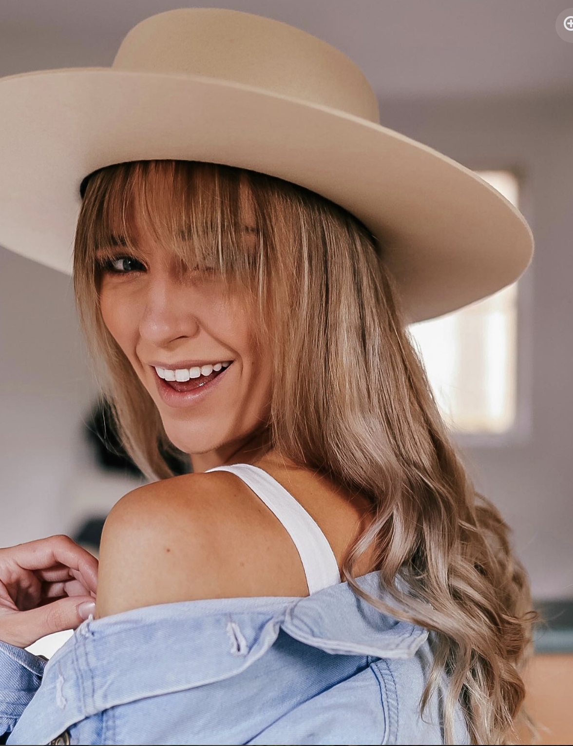 A woman smiling and looking over her shoulder, wearing a wide-brimmed hat and a blue denim jacket.