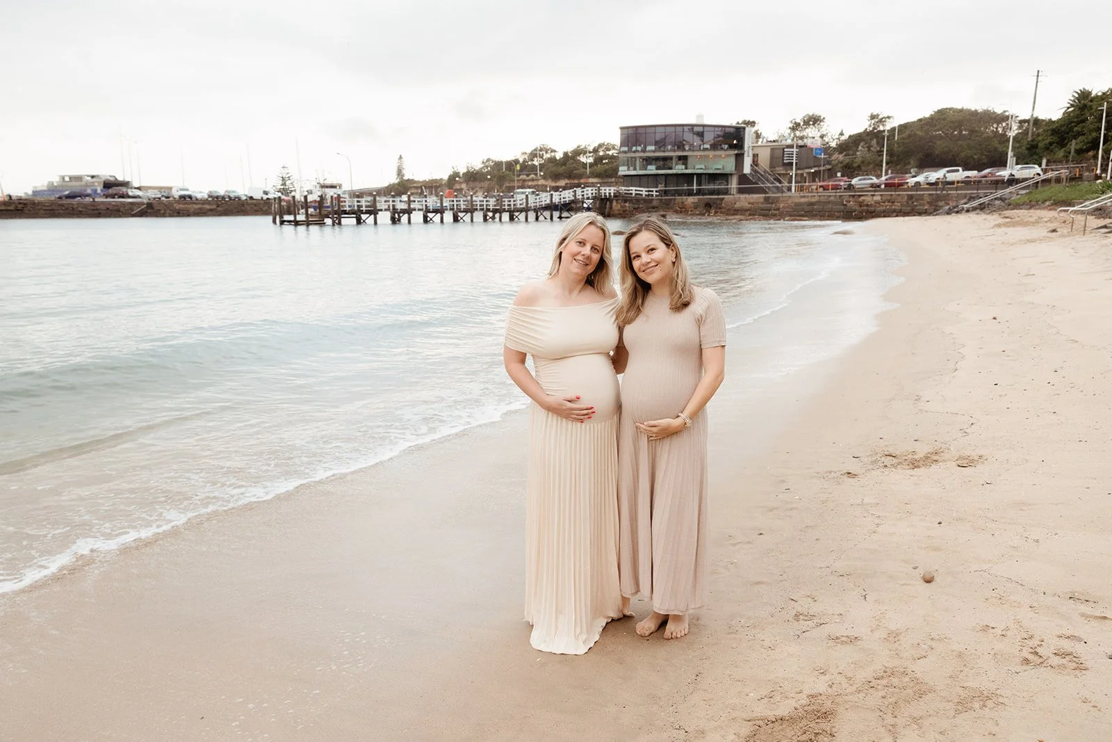 Two pregnant women standing on a sandy beach near the water, smiling, with buildings and a parking lot in the background.