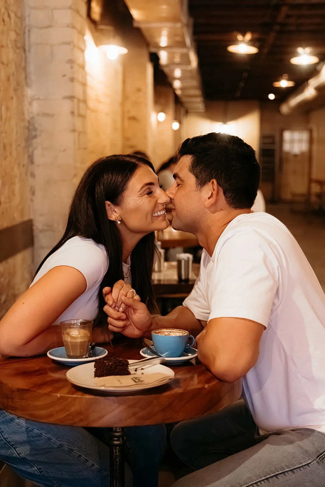 A couple sitting at a table in a cozy cafe, smiling and leaning in close, holding hands. The table has coffee drinks and a slice of chocolate cake.