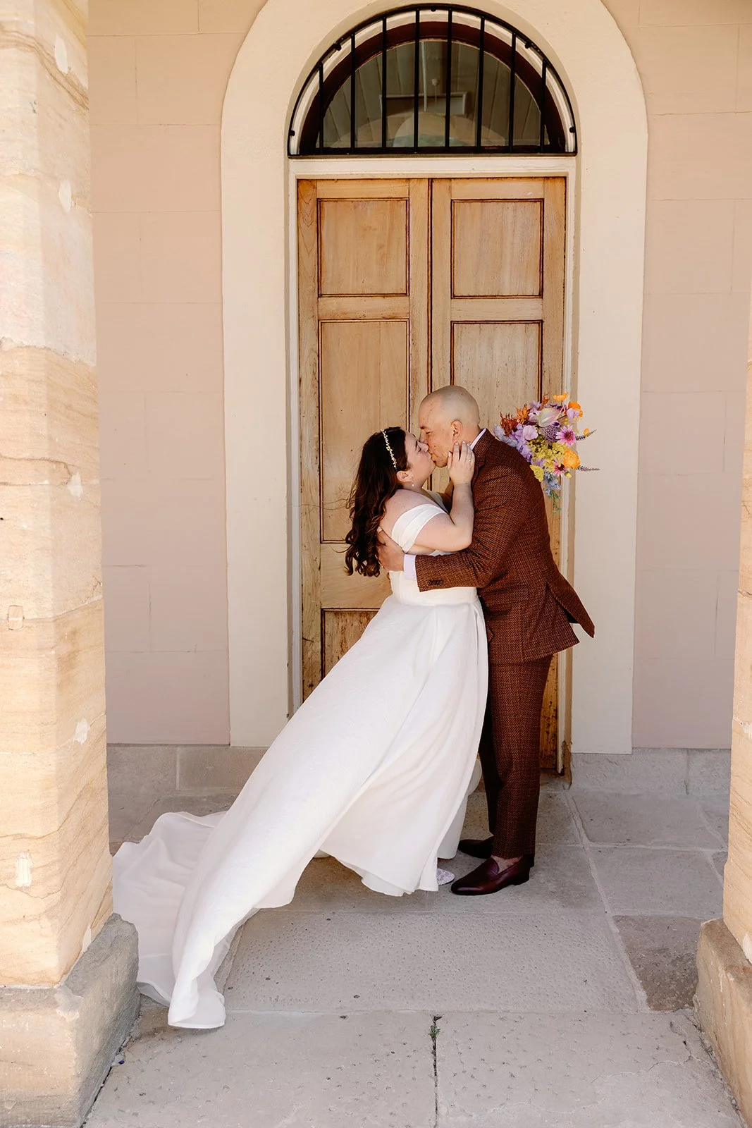 A bride and groom sharing a kiss in front of a wooden door with an arched window, the bride wearing a white wedding dress and the groom in a brown suit, holding a bouquet of colorful flowers.