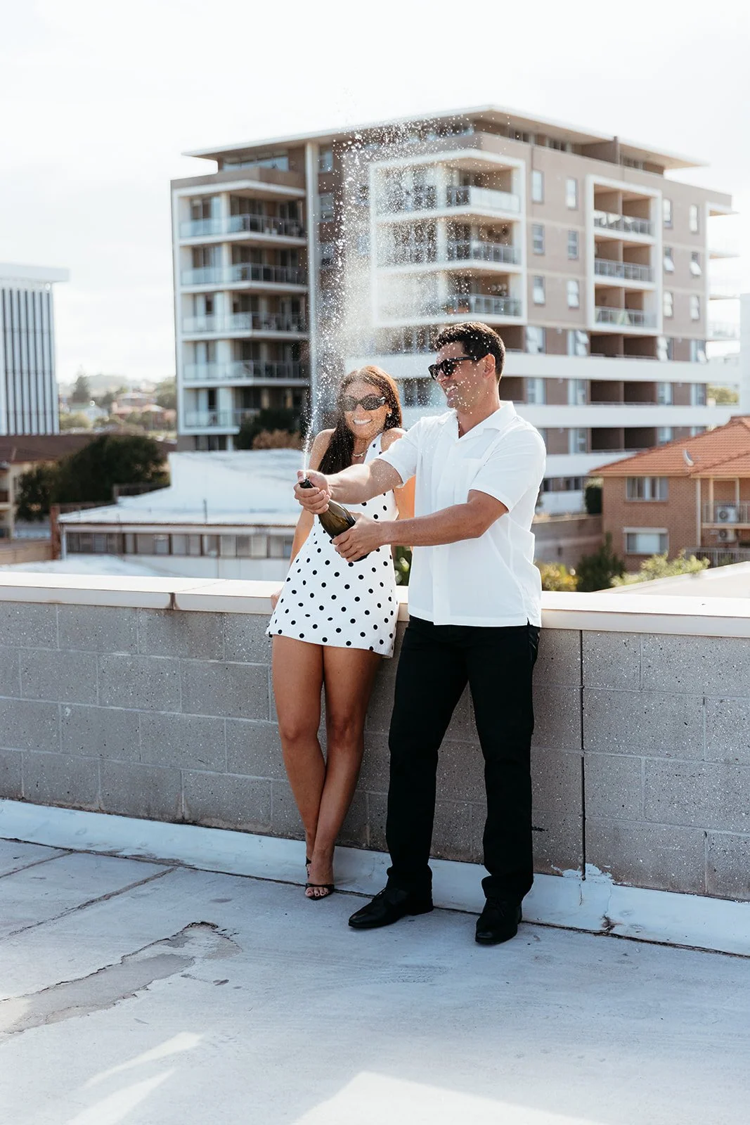 A man and woman celebrating on a rooftop with champagne, the man popping the cork and the woman smiling, both wearing sunglasses, with buildings in the background.