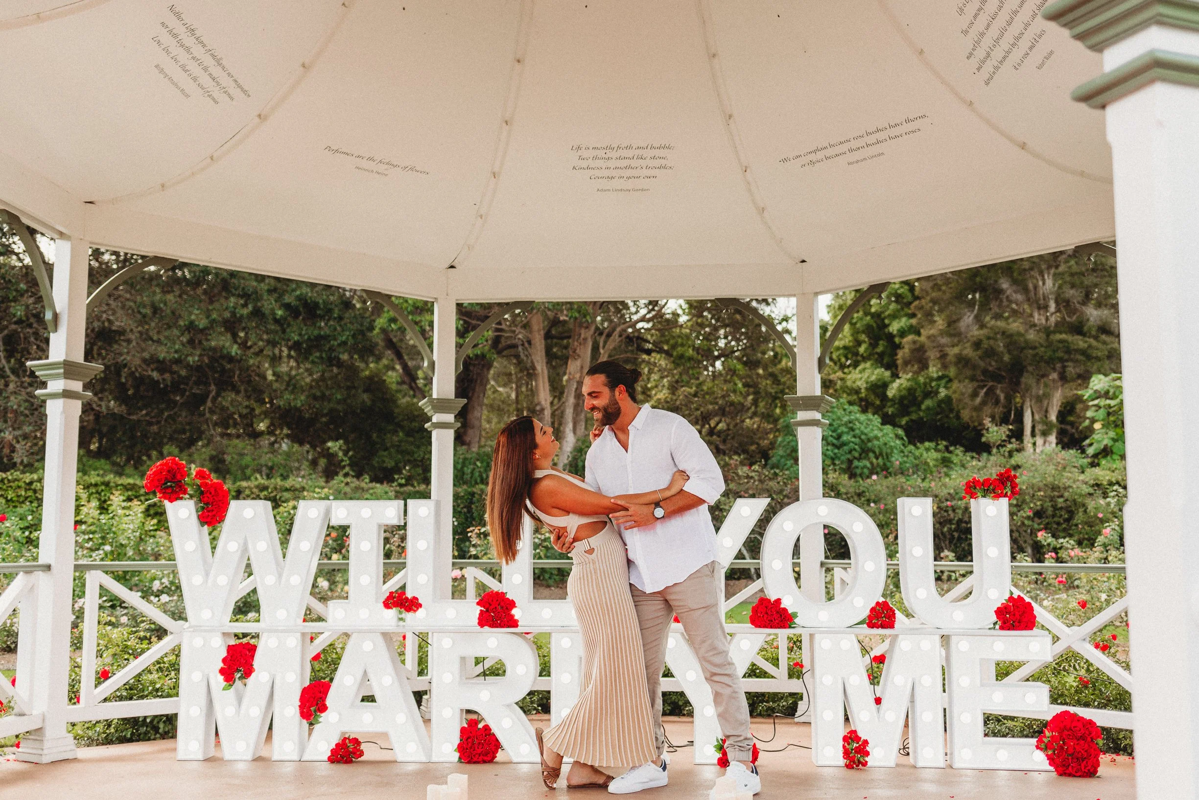 A couple dances together under a gazebo decorated with illuminated letters spelling 'WILL YOU MARRY ME' and red flowers in an outdoor garden setting.
