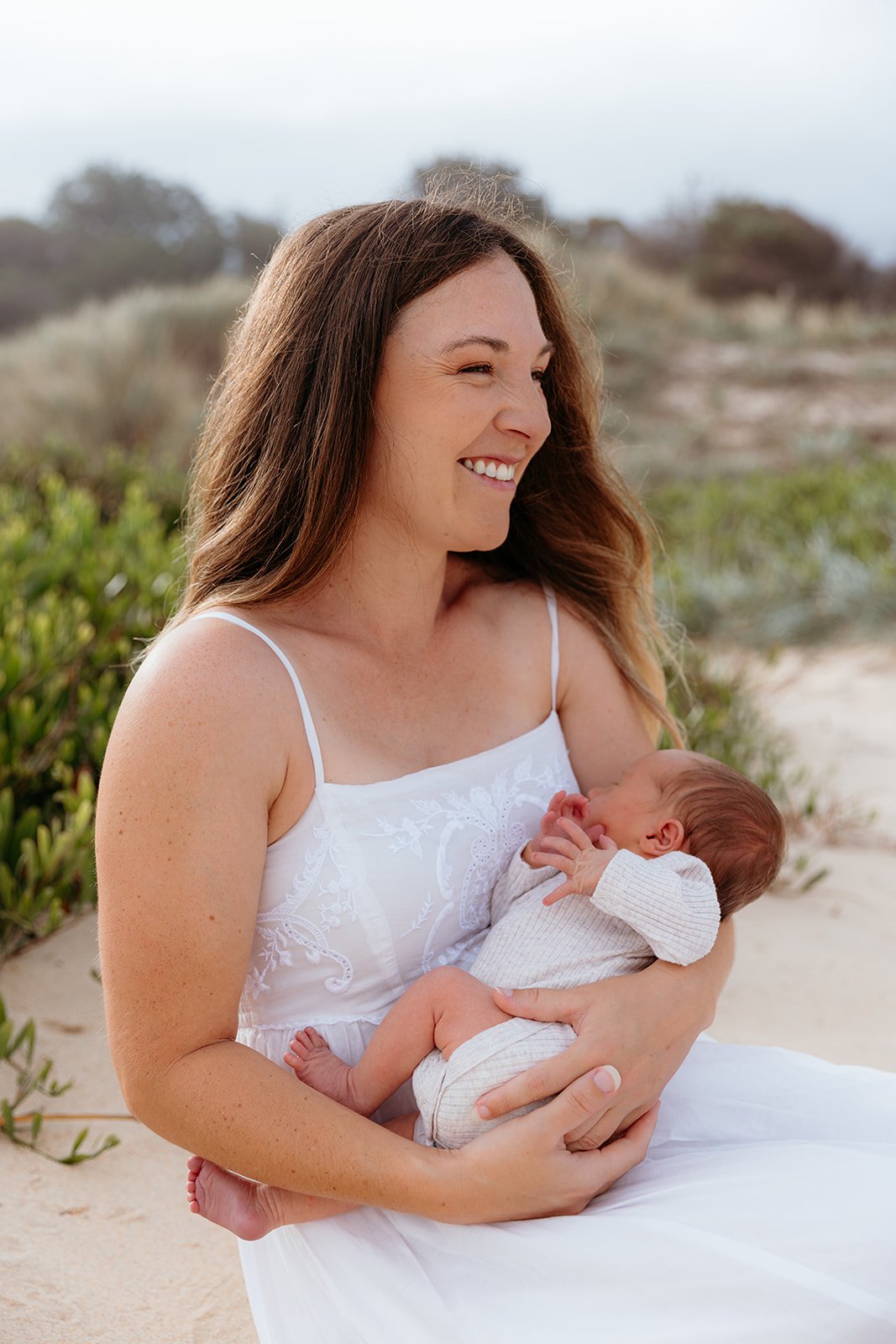 A woman in a white dress holding a newborn baby on a beach.