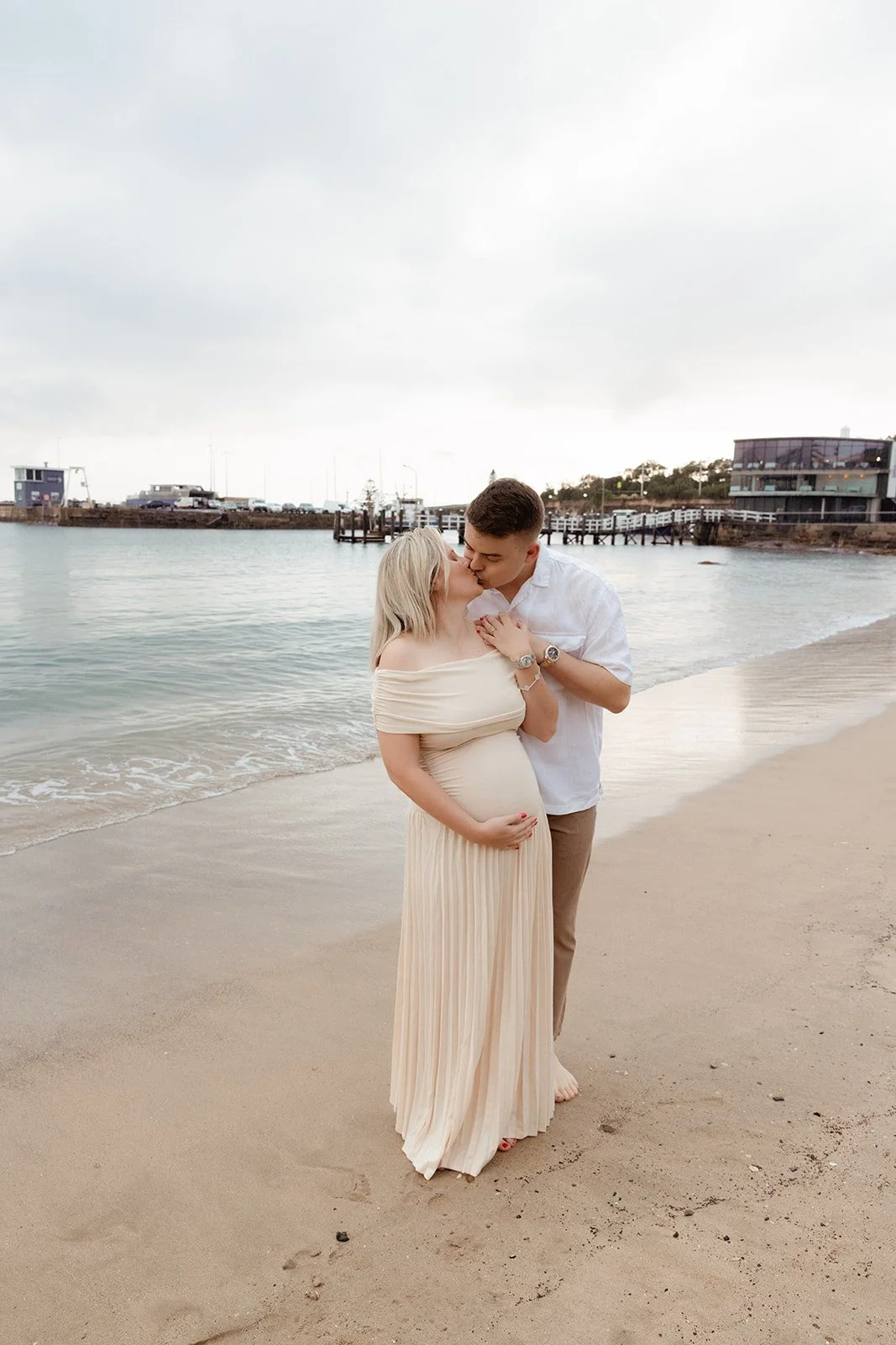 A couple kissing on the beach, the woman is pregnant and wearing a cream off-the-shoulder dress, and the man is dressed in a white shirt and beige pants.