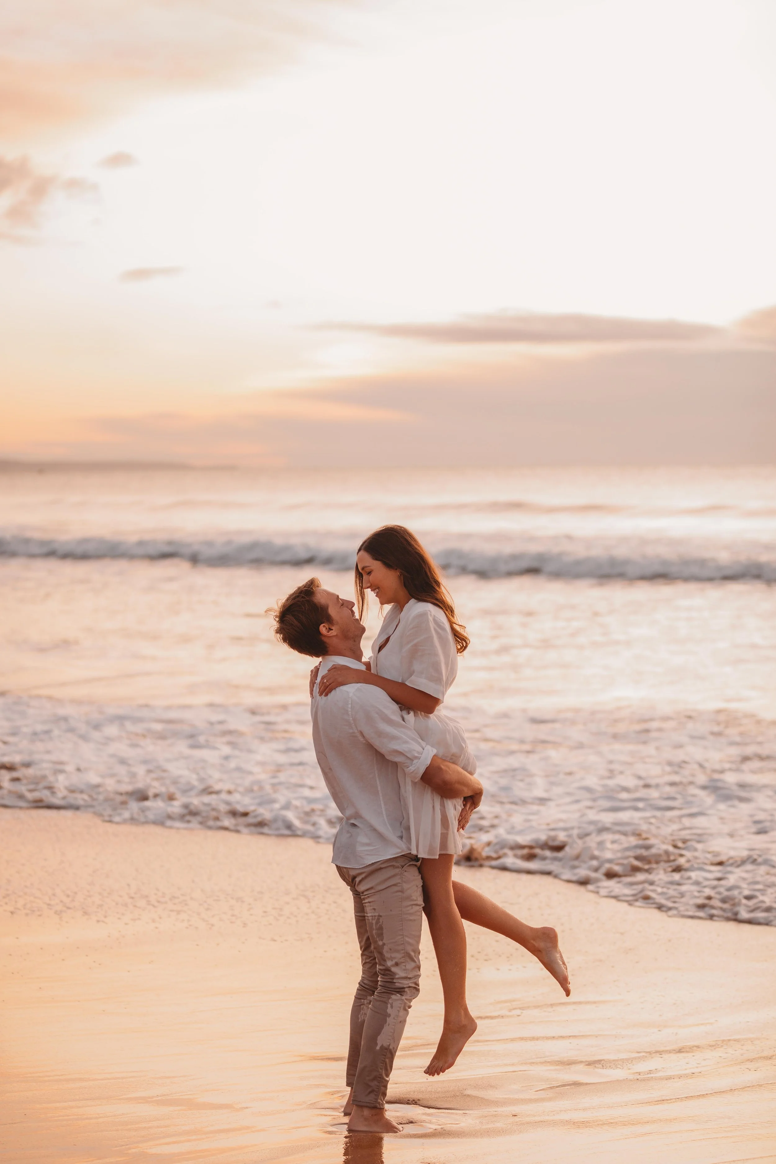 A man and a woman, both dressed in light-colored clothing, are embracing and smiling at each other on the beach during sunset. The man is lifting the woman off the sand, and they are near the shoreline with gentle waves in the background.