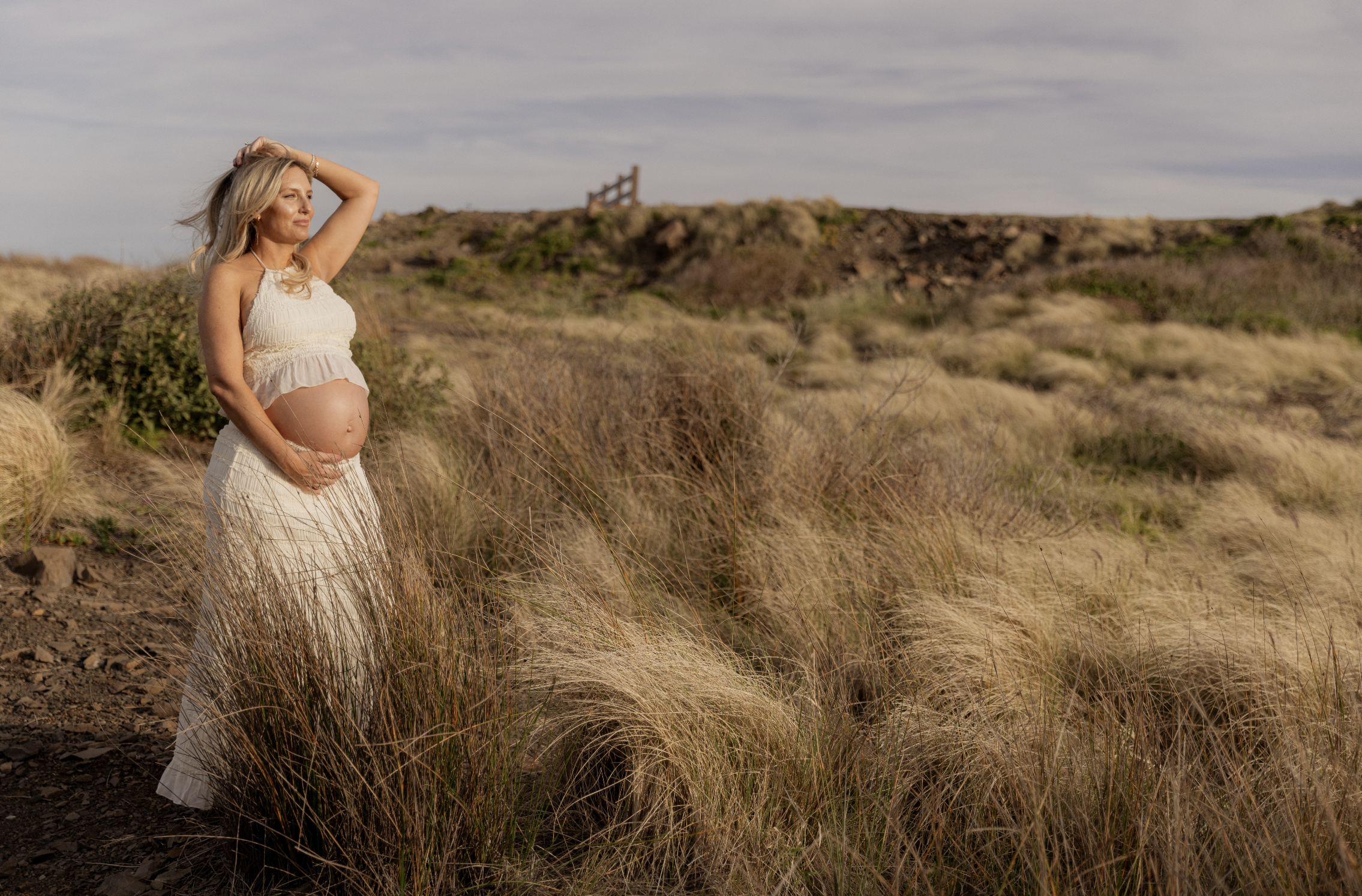 Pregnant woman in a cream-colored dress standing in a grassy field with shrubs and a hill in the background.