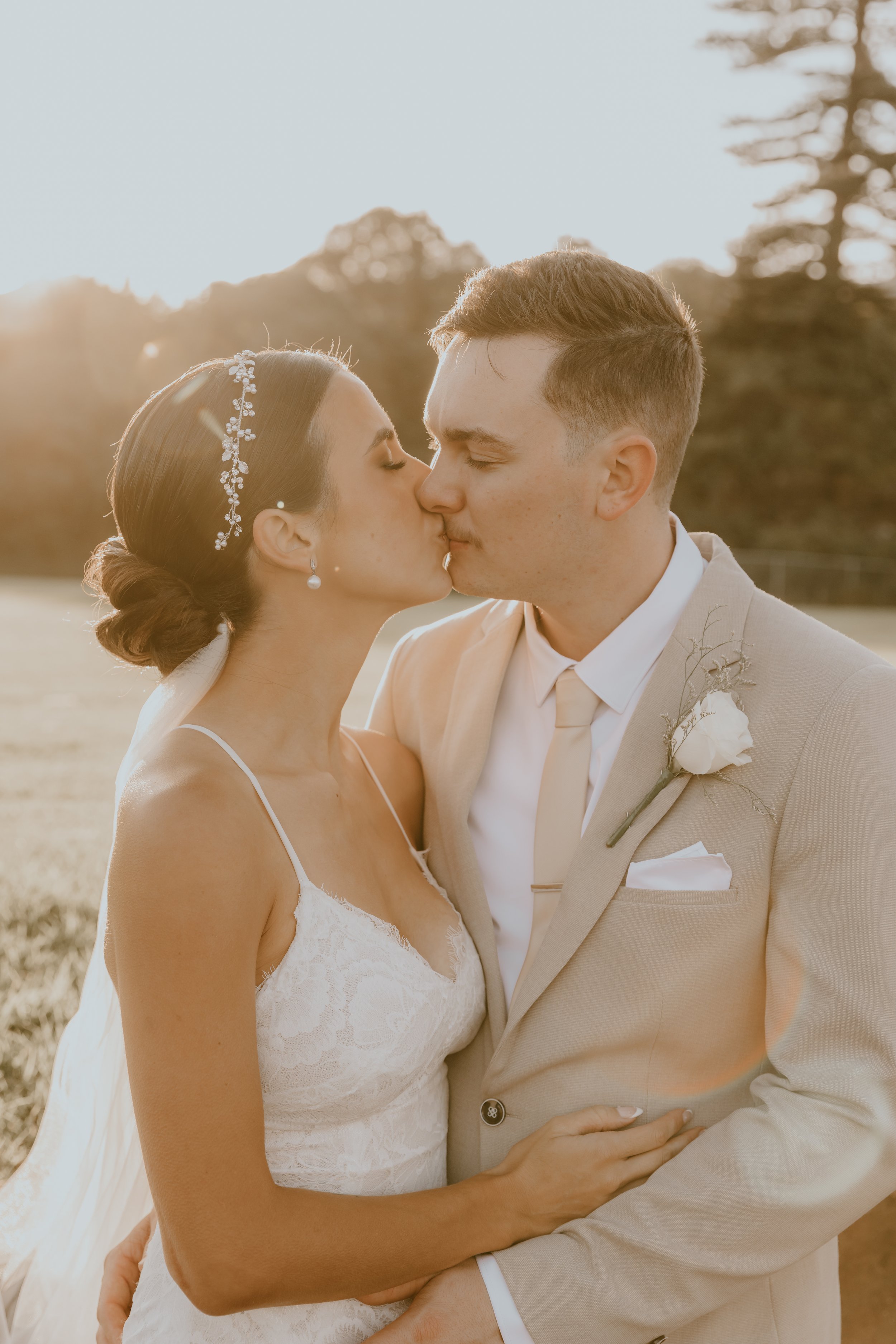 A bride and groom kiss outdoors during sunset, with trees in the background, dressed in wedding attire.