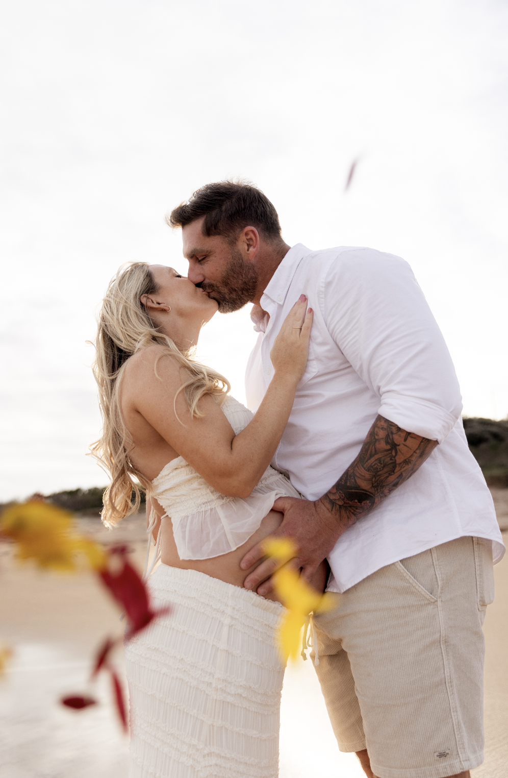 A couple sharing a kiss on a beach during daytime, with blurred flowers in the foreground.