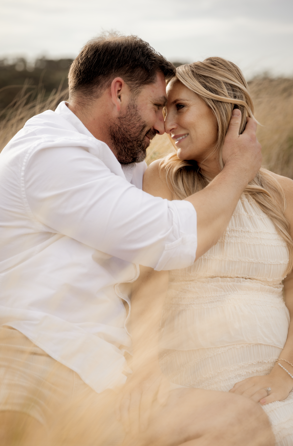 A romantic moment between a man and a woman outdoors, touching foreheads and smiling, with grassy fields and a cloudy sky in the background.