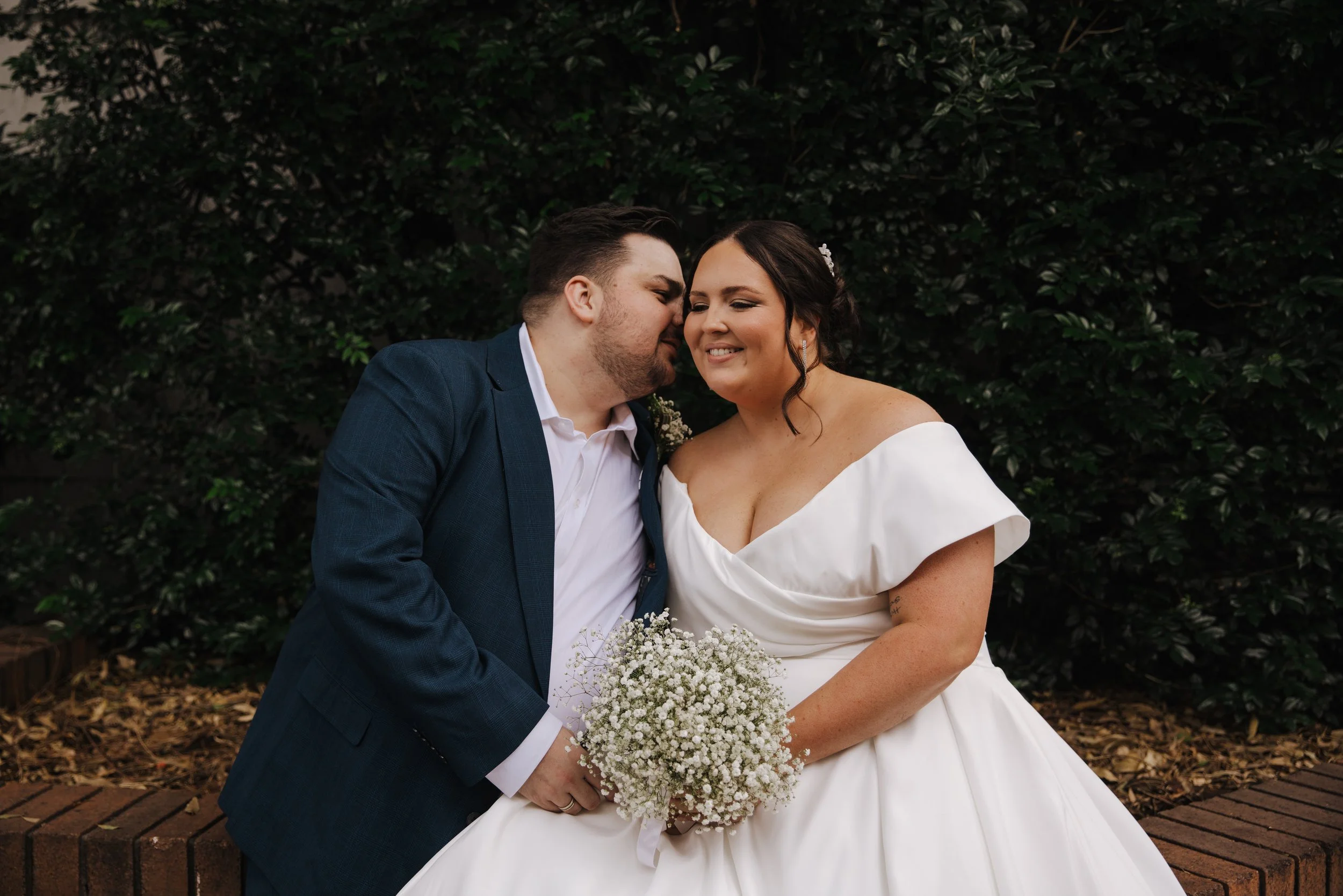 A newlywed couple sitting close together in front of a green leafy background. The bride is in a white off-shoulder wedding dress holding a white bouquet, smiling with closed eyes. The groom, in a dark blue suit with a white shirt, is leaning in towa