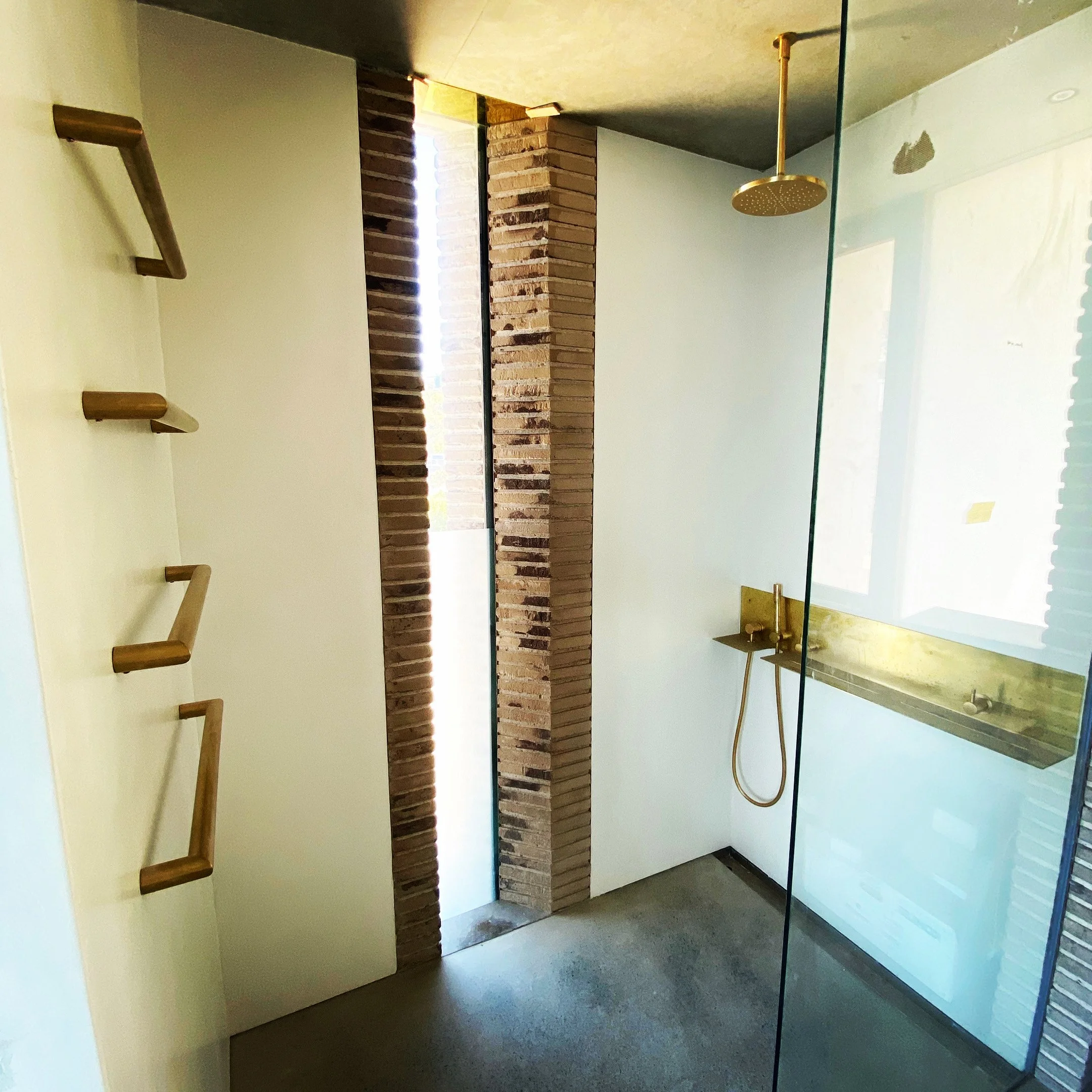Modern bathroom shower area with a glass wall, gold fixtures, and a built-in shelf, featuring brick pillar and wooden accents.