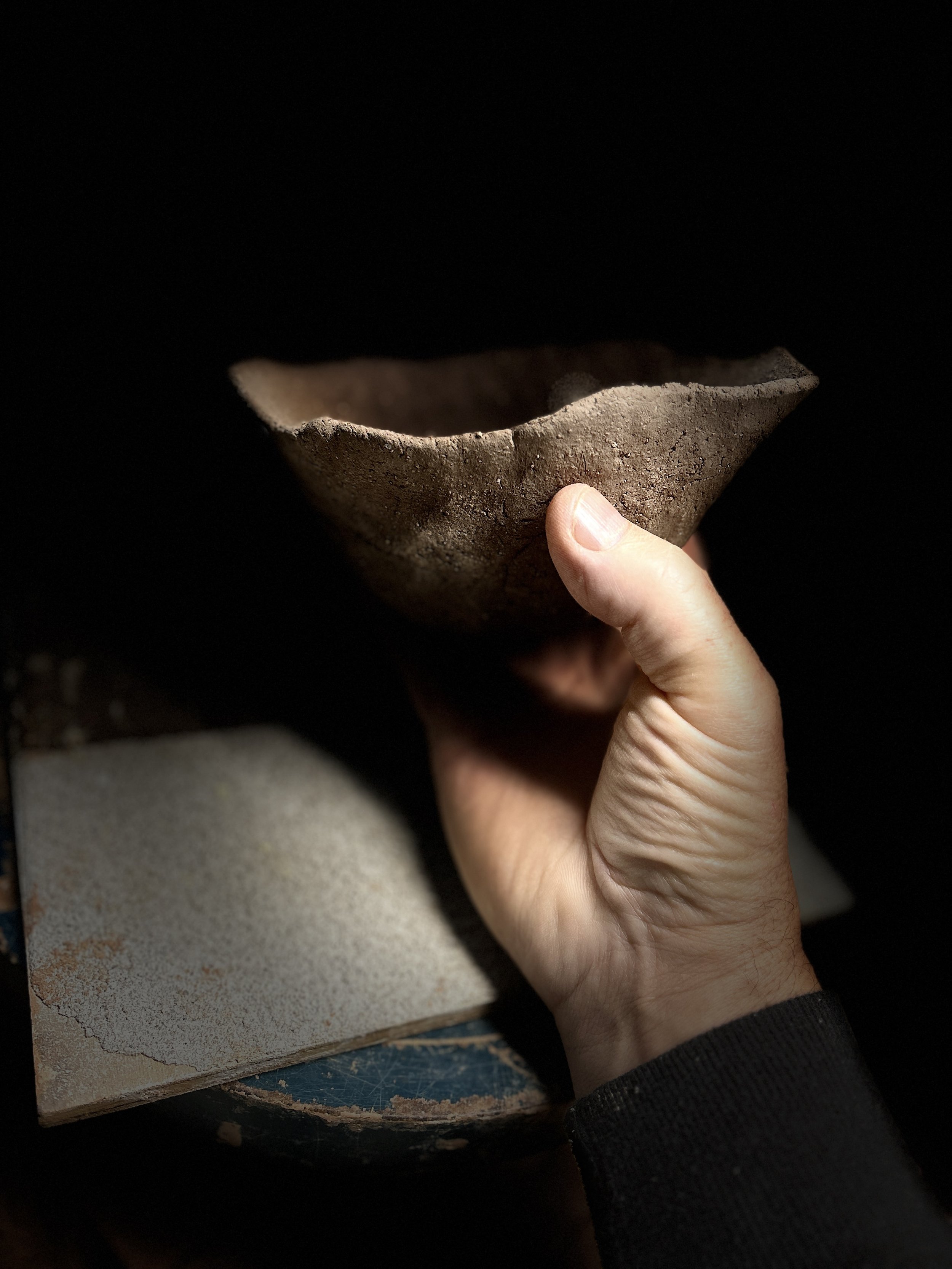 A person holding an ancient, broken clay bowl above a piece of pottery shard with a partially painted surface, with a dark background.