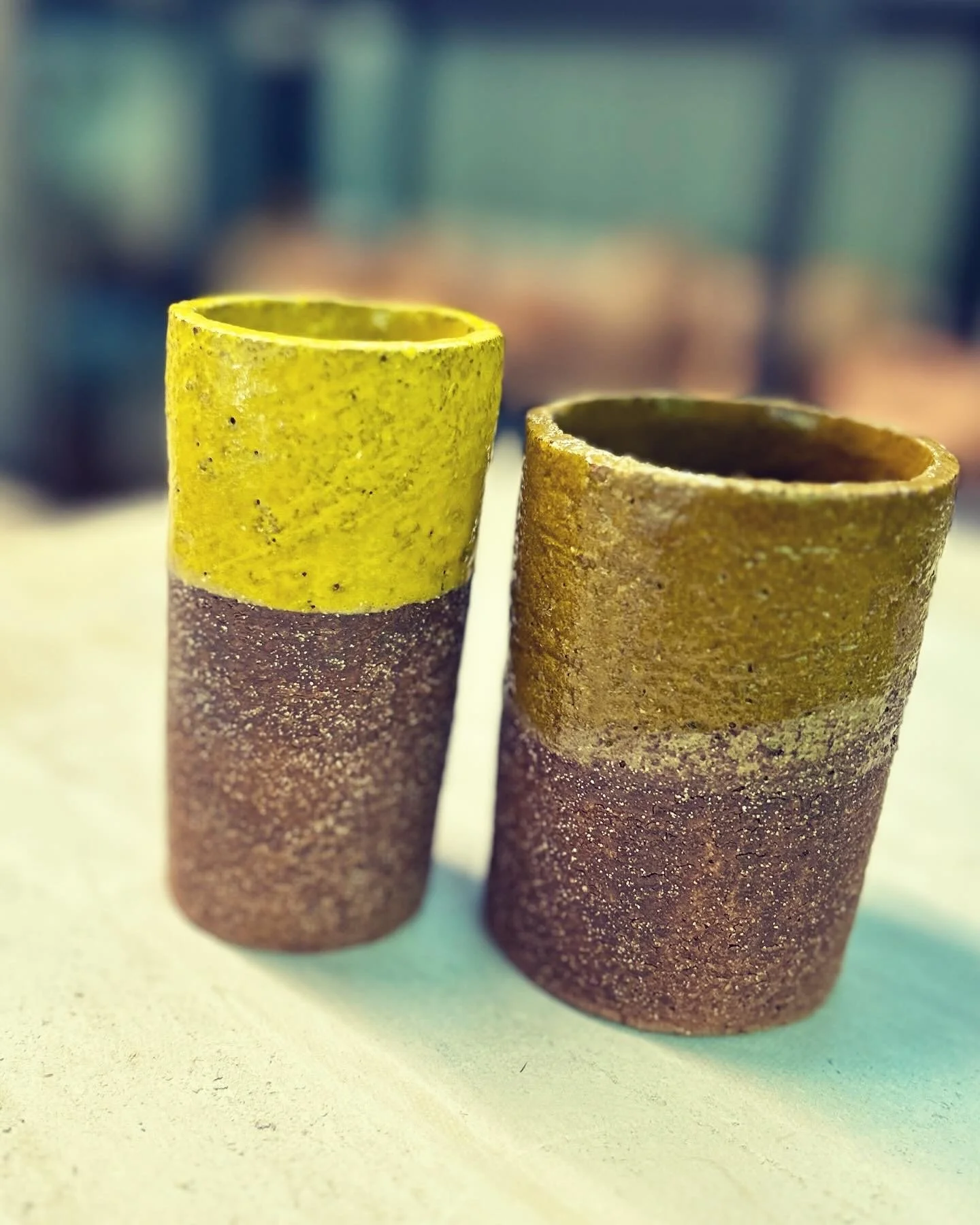 Two handmade ceramic cups with textured surfaces, one with a yellow top half and the other in a natural earthy color, placed on a light-colored surface with a blurred background.