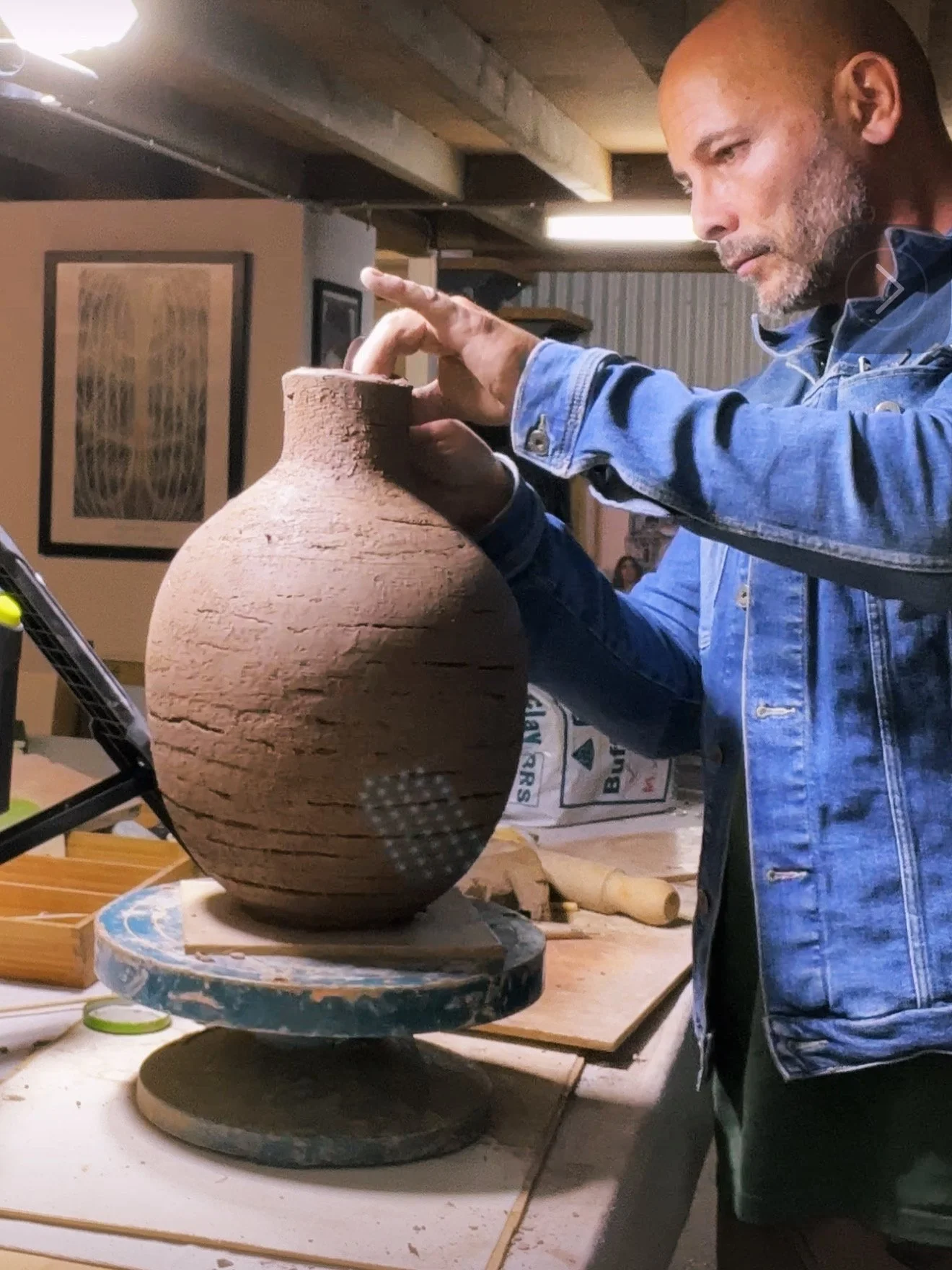 A man shaping a large clay vase on a pottery wheel in a workshop.