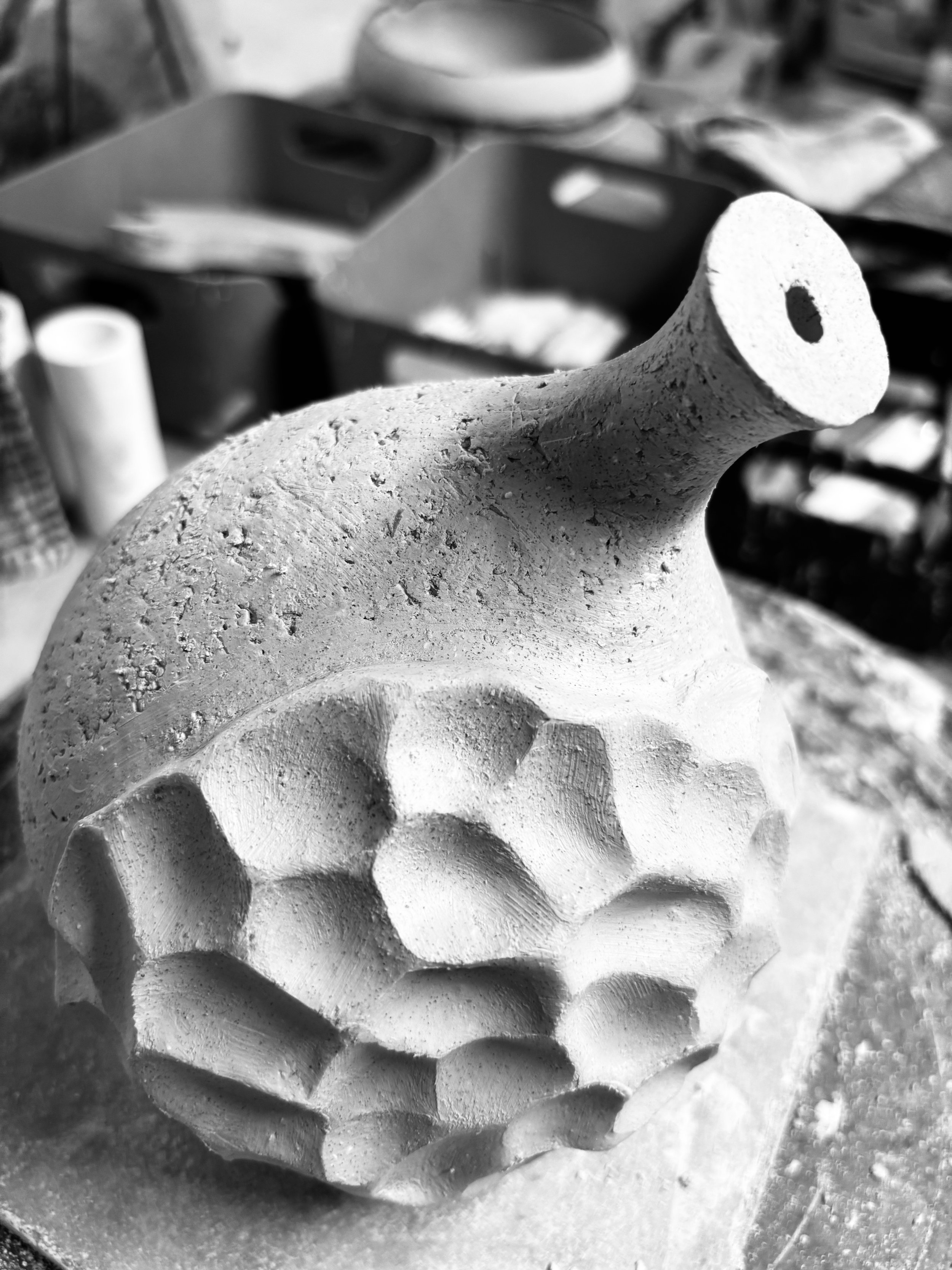 A ceramic object with a textured, patterned surface and a protruding tube, placed on a work surface with storage containers and other materials in the background.
