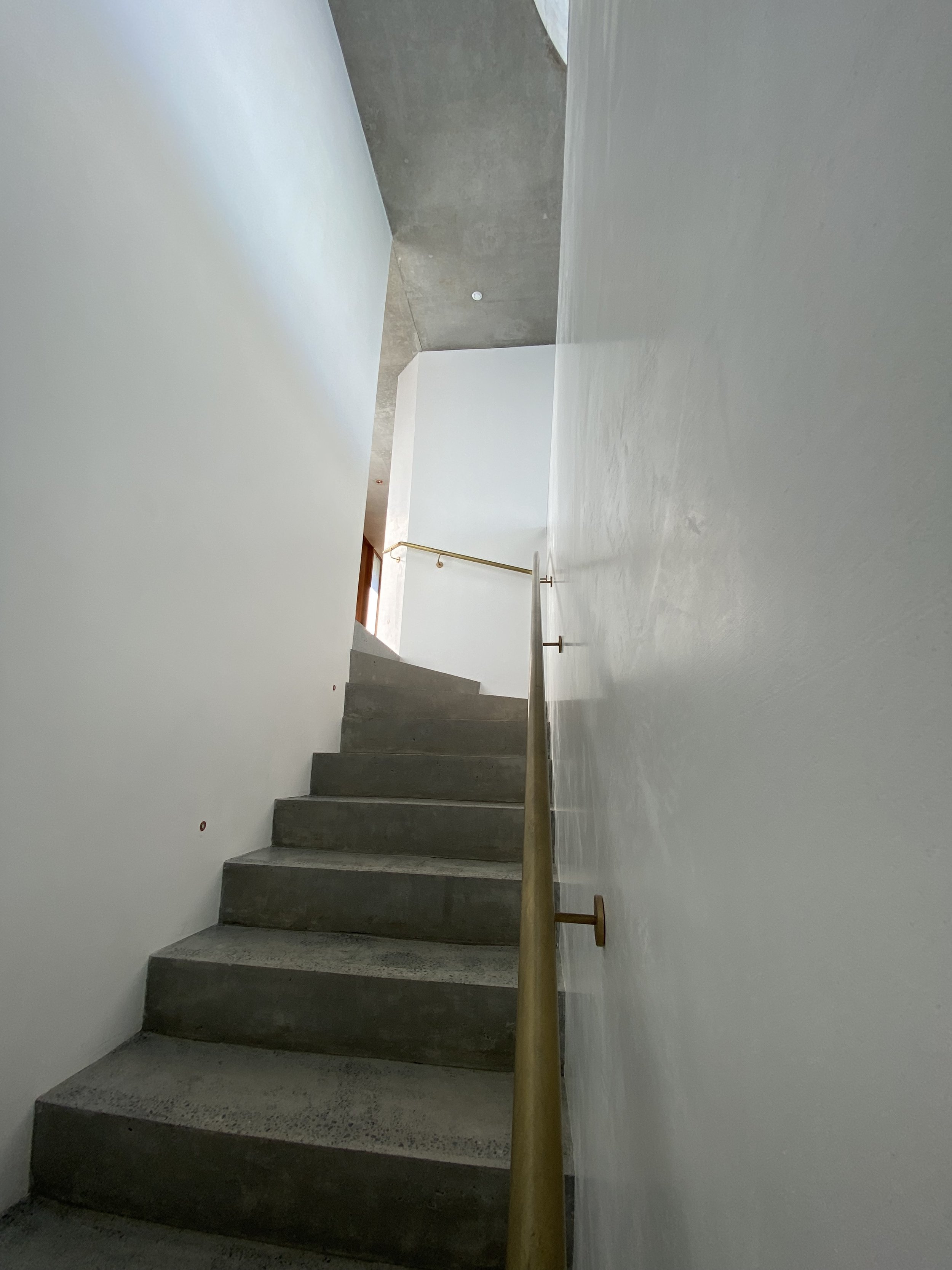 Photo of indoor staircase with concrete steps, white walls, a wooden handrail on the right side, and a small window near the top allowing natural light.