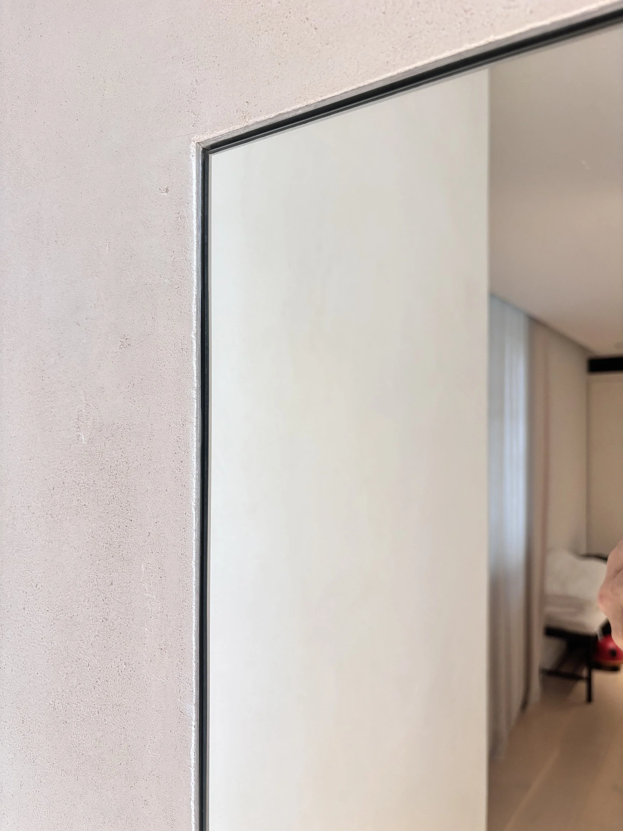 Close-up of a corner of a mirror on a wall in a hotel room, with part of the bed and a red item visible in the background.